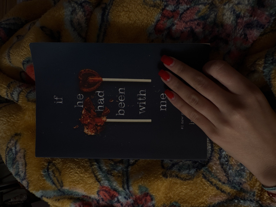 The image presents an overhead view of a dark-covered book titled "if he had been with me," resting on a yellow-orange patterned fuzzy blanket. The book's cover art depicts two lollipops with sticks: the left lollipop is a broken red heart, and the right is an intact red heart. A human left hand, displaying red manicured fingernails and a thin blue string bracelet with a red bead, is positioned across the lower right corner of the book. The environment appears to be an indoor setting in Los Angeles, United States of America, under dim lighting.
