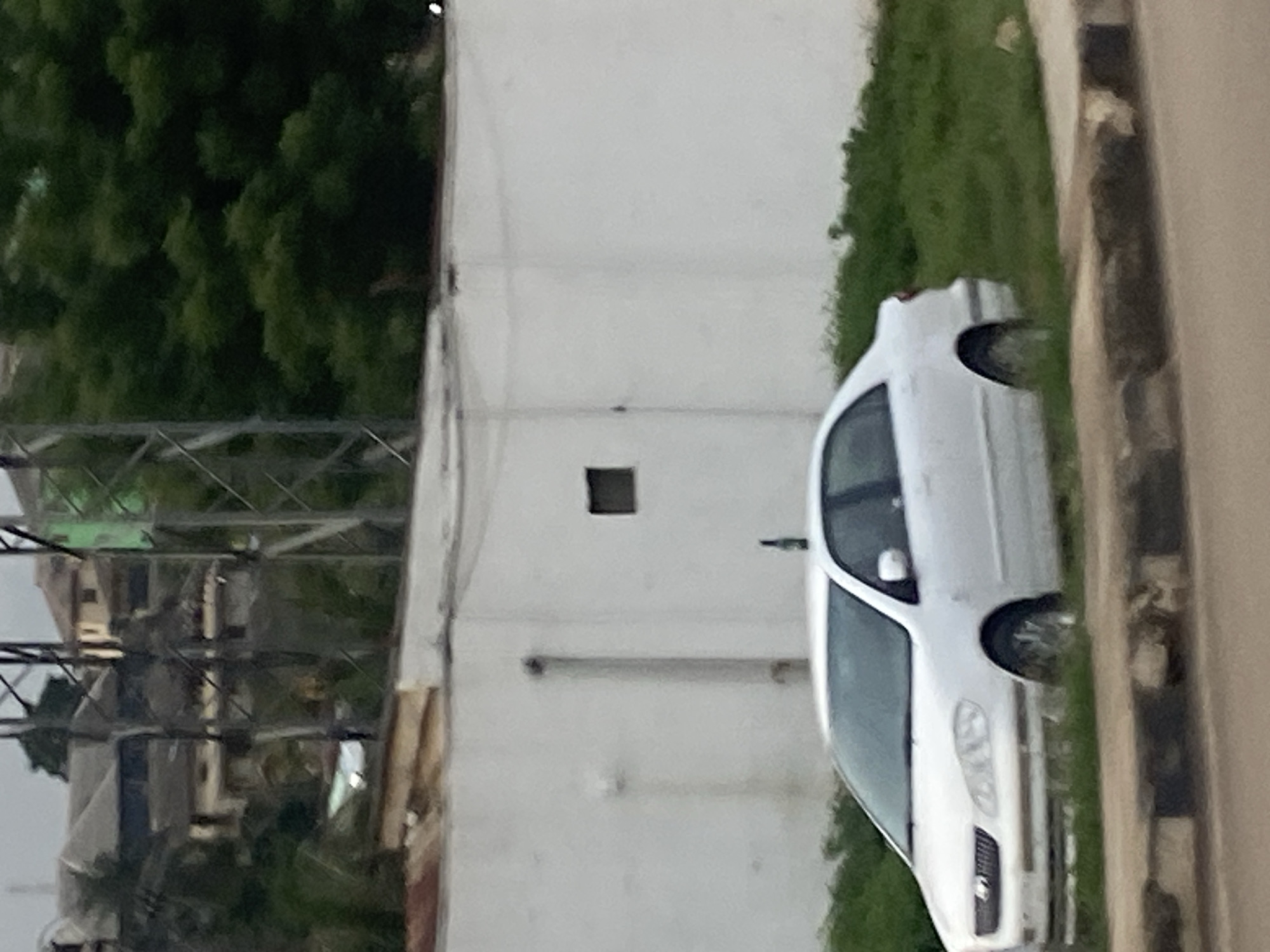 The image captures a slightly overcast day in Ikorodu, Nigeria. A white sedan is parked on the grassy shoulder of a road. The car is positioned diagonally, facing towards the left of the frame, with its front wheels slightly embedded in the overgrown grass. A dark object, possibly a bottle, is perched vertically on the car's roof antenna. Behind the car, a plain white wall stretches horizontally across the scene, interrupted by a small, dark rectangular window. In the background, several structures are visible, including buildings with grey roofs and some with tan-colored walls. Tall, green trees form a dense canopy to the right of the frame, and a metal communication tower with a lattice structure stands prominently near the center-left of the image. The road in the foreground has a textured curb made of concrete and darker stones.