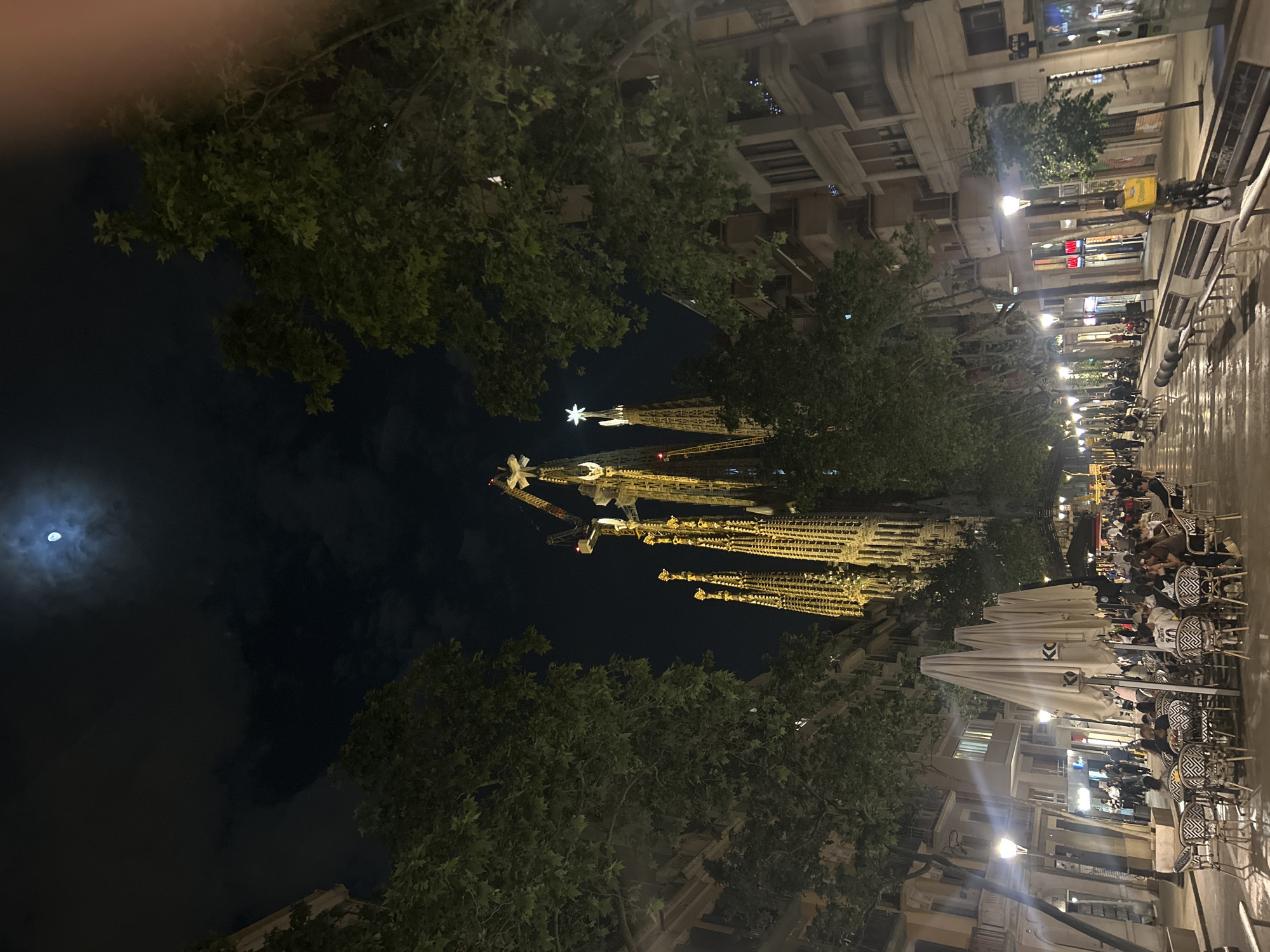 This is a nighttime view of the Sagrada Familia in Barcelona, Spain, taken from street level. The basilica's illuminated spires rise prominently in the center of the frame, framed by trees on both sides. In the upper left, a visible moon is partially obscured by clouds. To the right, the street is lined with cafes, with tables and chairs occupied by people. Streetlights cast a warm glow on the sidewalk and buildings. The scene suggests a lively evening atmosphere with both the iconic architectural landmark and active street life.