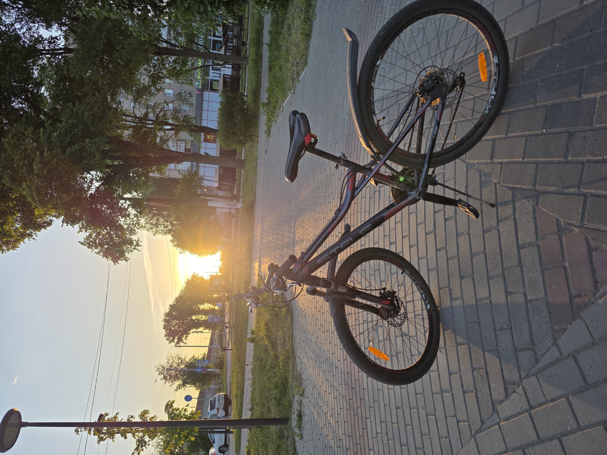 The image shows a "Stels" brand bicycle parked on a gray brick sidewalk, possibly in Chaikovsky, Russia. The bicycle faces the setting sun, with trees and buildings visible in the background. A road with cars is to the left. The bike is upright with both wheels on the ground. The rear wheel has a mudguard. The lighting is warm due to the sunset.