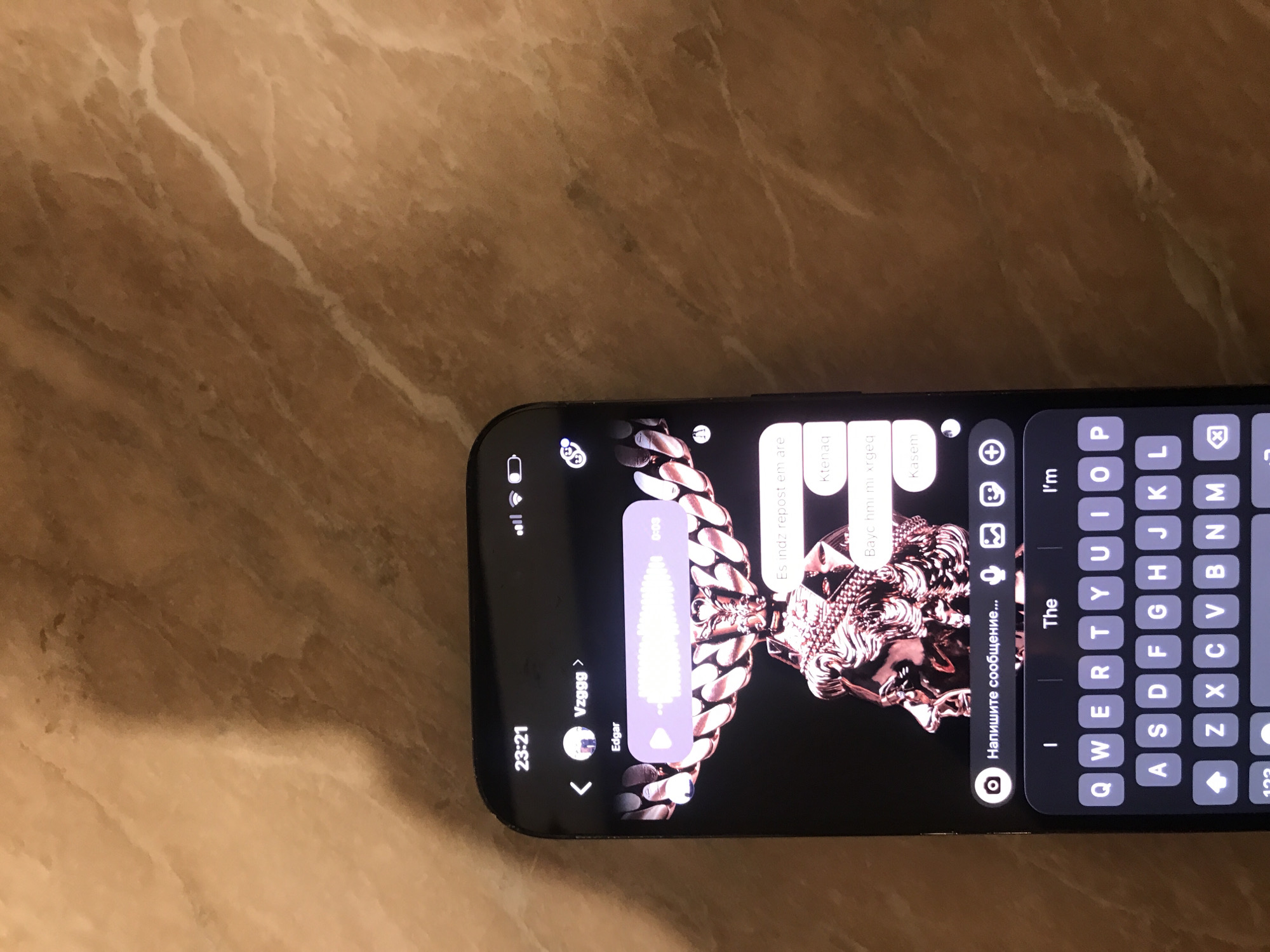 A mobile phone is lying on a surface with a marble-like pattern. The phone screen displays an open messaging application. The sender's name is "Vzggg" and their real name is "Edgar". There is a short voice recording in the chat. The content of the messages exchanged in the chat are written in a language other than English. The keyboard of the phone is visible at the bottom, ready for typing. The time displayed on the phone is "23:21". The image lacks context that can identify the specific location in Yerevan, Armenia.