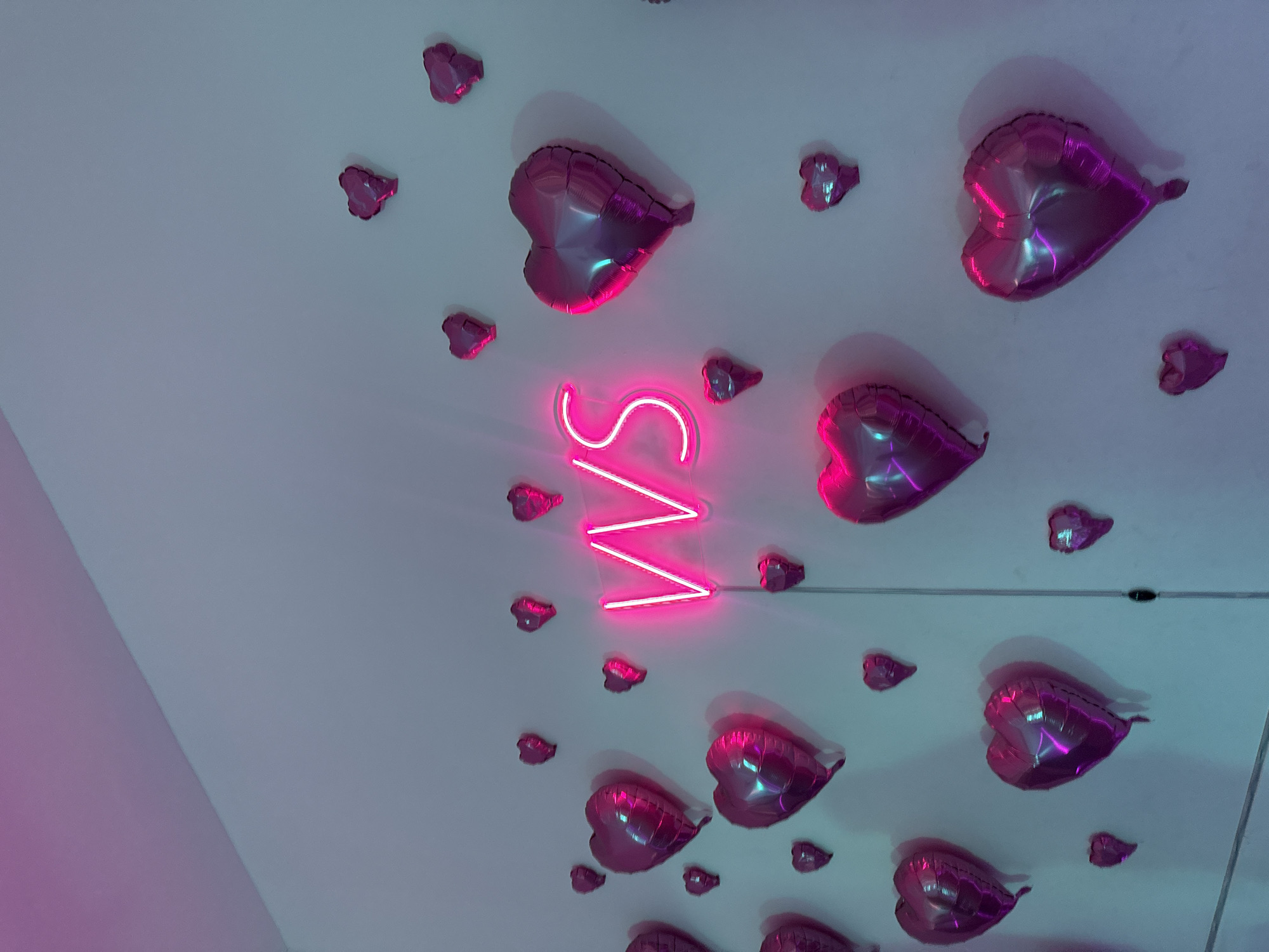 An indoor, low-light image captured at an angle showcases a wall or ceiling adorned with numerous metallic heart-shaped balloons and a glowing neon sign. The background surface is a light, cool grey or white, with areas reflecting purple and blue ambient light.
The balloons, varying in size, are a vibrant pink or fuchsia, with glossy, reflective surfaces that catch the light, showing bright highlights and darker shadows. They are scattered across the surface, giving a decorative and celebratory feel. Some balloons cast faint, soft shadows on the wall.
Centrally positioned is a bright pink neon sign, emitting a strong glow that illuminates its surroundings. The sign clearly spells out the capital letters "VVS". The light from the neon sign casts a radiant pink hue onto nearby balloons and the background.
No people are visible, and no specific activity is taking place. The arrangement of hearts and the neon sign suggest a themed event or celebration, possibly romantic in nature like Valentine's Day, an anniversary, or a lively party atmosphere. The setting is likely an event space or decorated room in Saint Petersburg, Russia. The overall mood is festive and modern, with a strong emphasis on pink and magenta tones against a cooler backdrop.