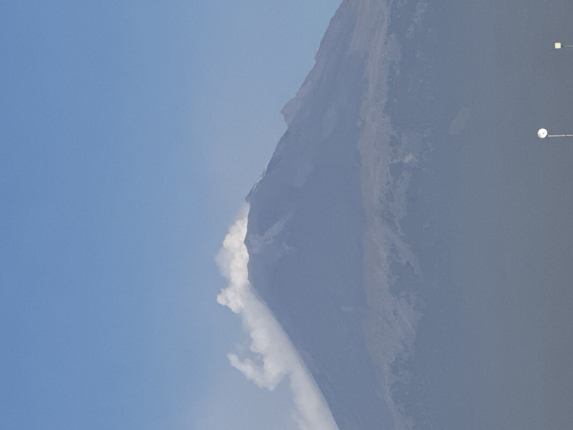 The image captures a majestic mountain or volcano under a clear, bright blue sky during daylight hours. The mountain dominates the right side of the frame, exhibiting a rugged, rocky profile in shades of grey and brown, with some areas appearing more textured, possibly indicating rock formations or sparse vegetation. A prominent white plume of vapor or smoke is actively emerging from the upper slopes of the mountain, extending downwards along its side, resembling a flowing cloud or a cascade. This significant plume suggests ongoing volcanic activity, such as steam or ash emissions.
The sky on the left side of the image is consistently clear blue, contrasting with the hazy appearance around parts of the mountain, which could be atmospheric conditions or distance. In the far upper right corner, two faint, light-colored points on vertical structures are barely visible, possibly streetlights or communication poles, indicating some human presence or infrastructure in the wider landscape. The scene conveys a sense of both natural grandeur and dynamic geological activity. Given the provided location context of San Francisco Ocotlán, Mexico, this is highly likely a depiction of the Popocatépetl volcano during one of its eruptive or degassing phases. There is no visible text in the image.