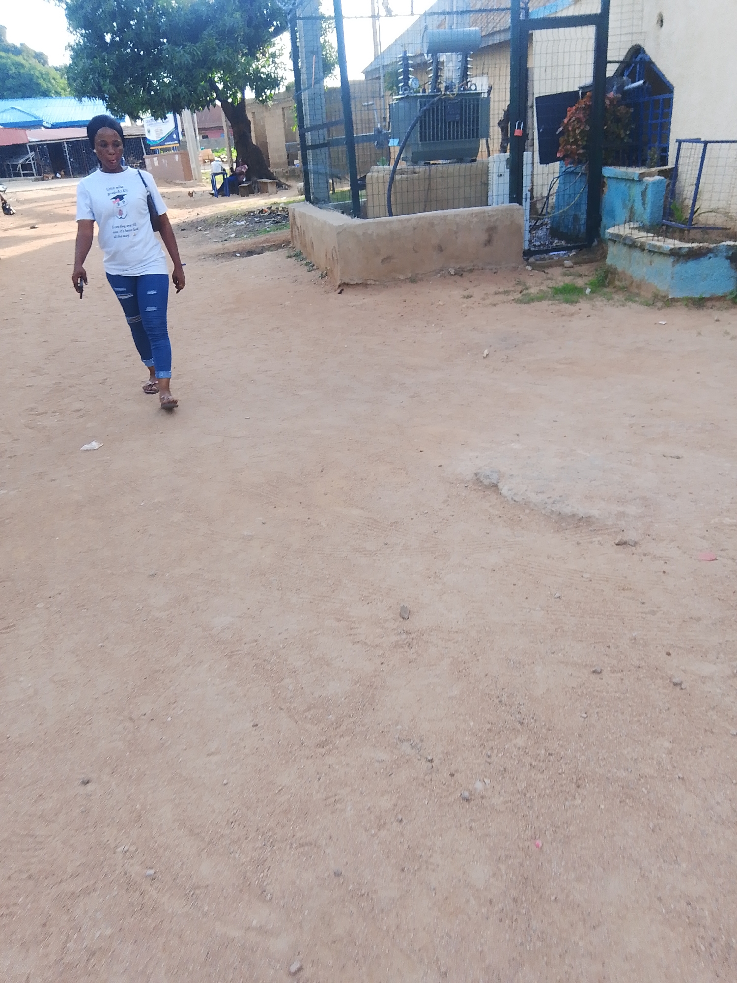 The image shows a woman walking on a dirt road in Jalingo, Nigeria. She is wearing a white t-shirt with text and graphics, blue ripped jeans, and sandals. A dark handbag is slung over her left shoulder. The background includes a large tree, a transformer, a fenced area, and some buildings with blue roofs. The ground is dusty and uneven, with some loose stones and debris. In the distance, other people and a motorcycle can be seen. The lighting suggests it is daytime, possibly late afternoon, with the sun casting shadows. The overall scene depicts an outdoor, casual setting in a semi-urban or rural area. The text on the woman's t-shirt is partially visible and appears to be in English, possibly a motivational quote or brand name. The setting suggests a warm climate, typical of Nigeria.