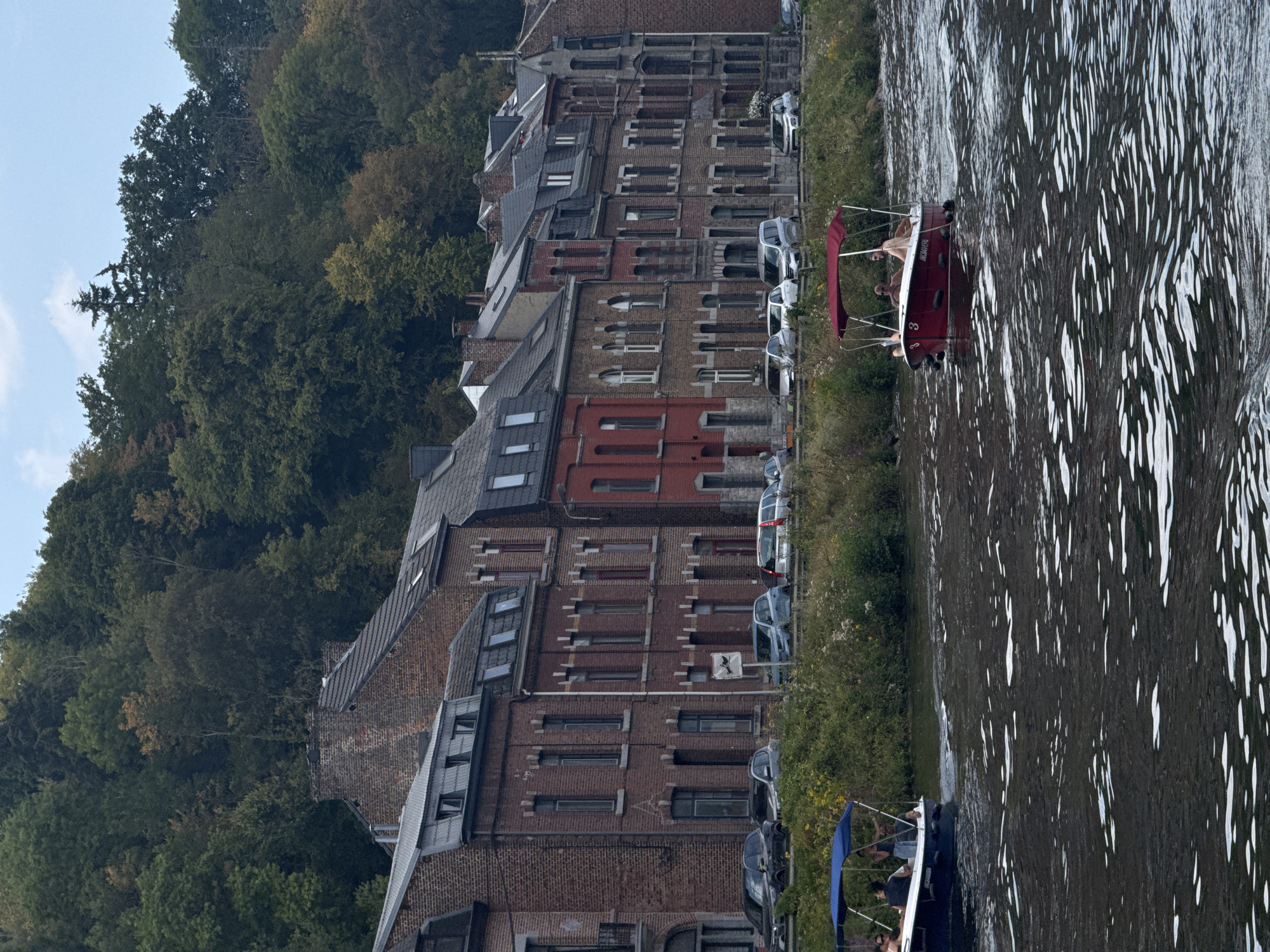 The image depicts a scene along the Meuse River in Dinant, Belgium. On the left, a blue-canopied boat carries at least three individuals, one of whom appears to be at the helm. Further to the right, a red-canopied boat contains two individuals. The river's surface is disturbed, indicating movement from the boats.
The background features a row of historic brick buildings lining the riverbank. These buildings have multiple stories, red brick facades, and varied architectural details including windows and rooflines. A tree-covered hillside rises behind the buildings, displaying autumnal foliage with greens, browns, and hints of yellow. Along the riverbank in front of the buildings, a grassy area with wildflowers is visible, and a line of parked cars is present. A traffic sign is also visible near the parked cars. The sky above is partly cloudy.