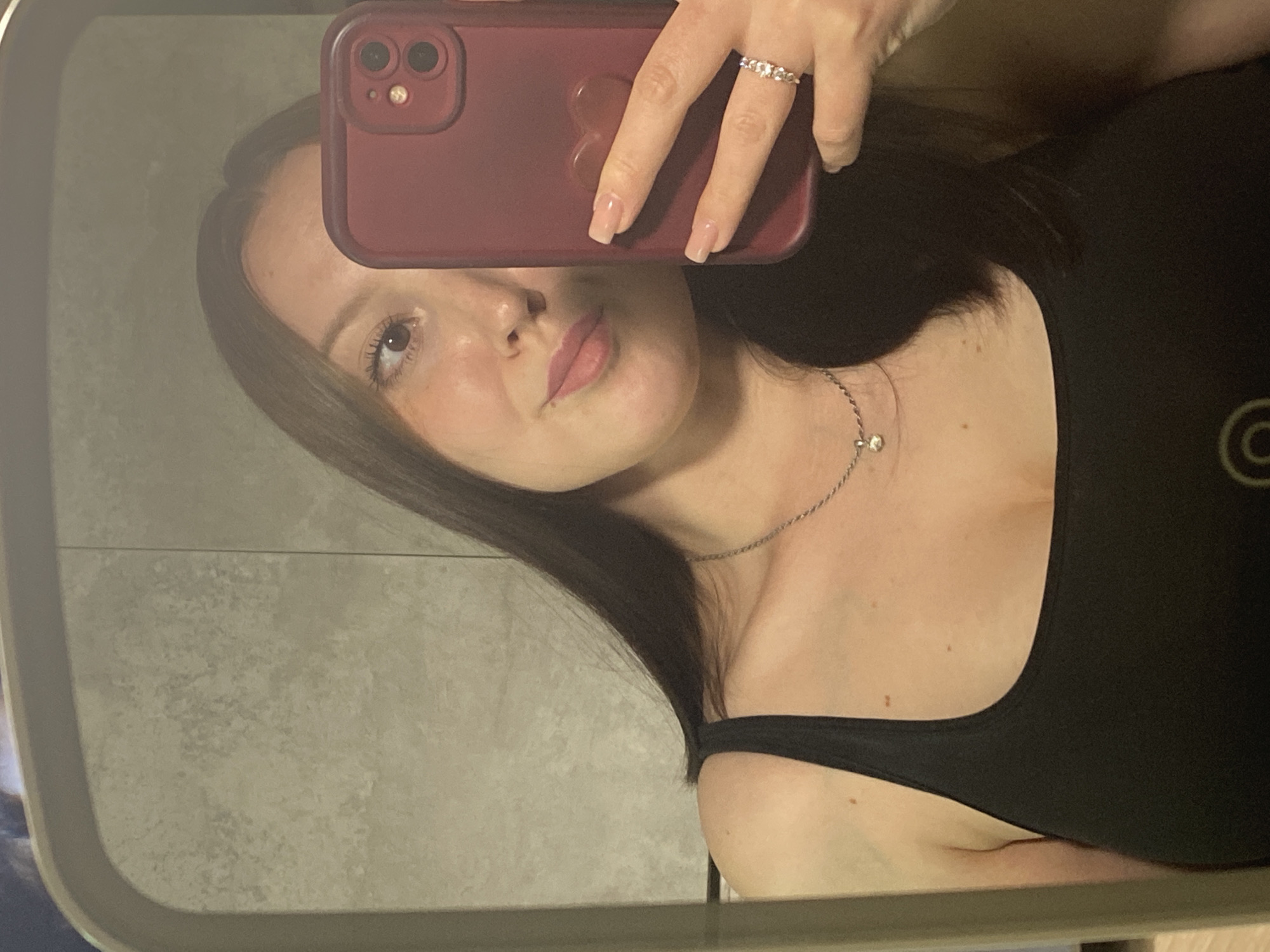 A woman is taking a selfie in a mirror. She is holding a burgundy-colored iPhone in her left hand, partially obscuring her face. She has long, dark brown hair and is wearing a black tank top and a silver necklace. She is also wearing a ring on her left hand. The background appears to be a bathroom with grey tiled walls and a mirror frame. The location is unknown and cannot be inferred from the image. The image does not depict the city of Chaikovsky, Russia.