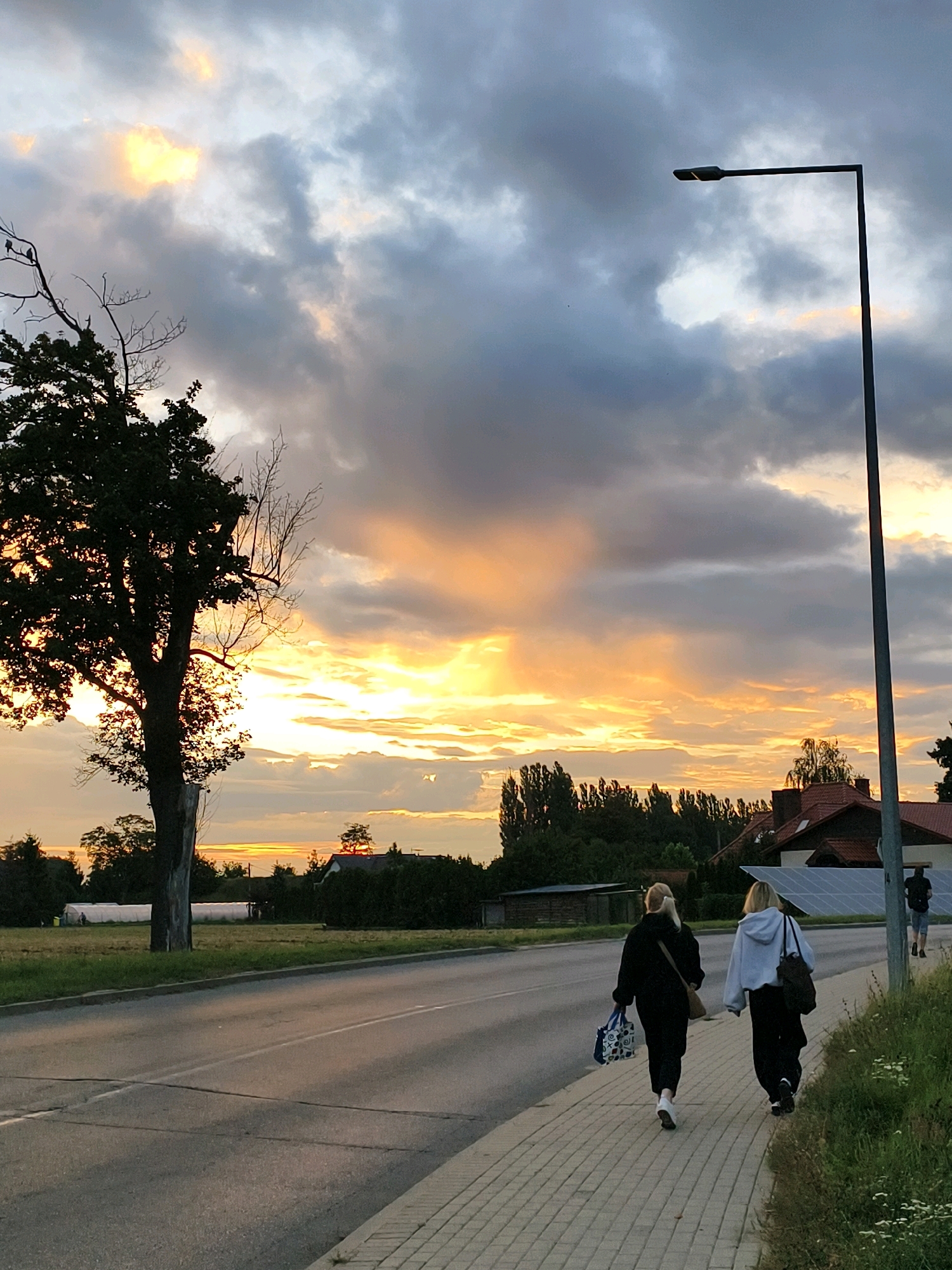 The image shows a street scene at dusk in Tczew, Poland, with a sunset casting warm light on the clouds. Two young women, one in a black hoodie and the other in a grey hoodie, walk away from the camera on a brick sidewalk. They are carrying bags. A tall lamppost stands on the right side of the sidewalk. To the left of the sidewalk, there is a road with cars visible in the distance. Behind the road, there is a large tree silhouetted against the sky, with some houses and trees further in the background. The sky is filled with dramatic clouds tinged with orange and yellow from the setting sun.