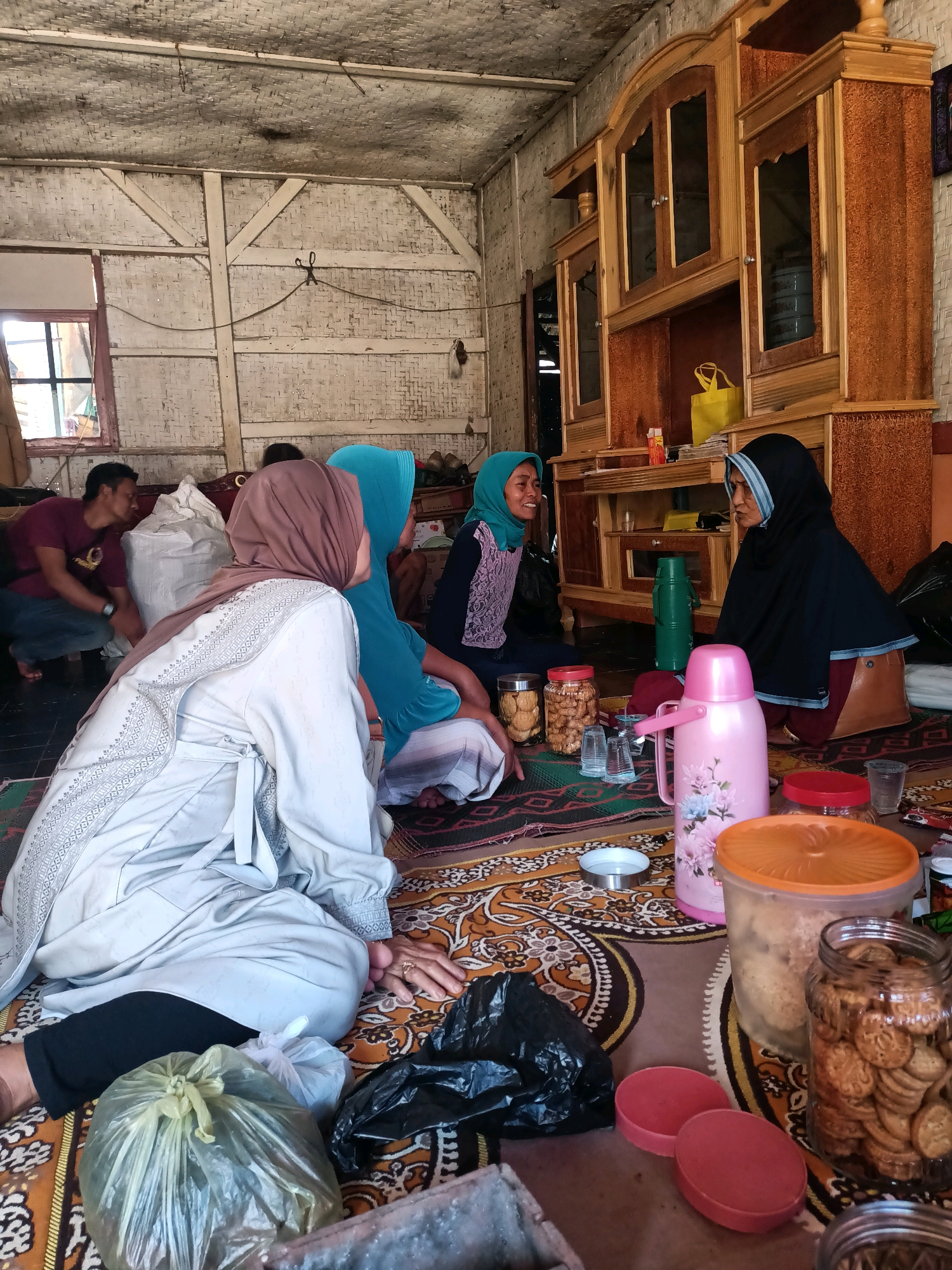 In Citamba, Indonesia, a group of four individuals, three women and one man, are gathered on a patterned rug within a rustic interior. The room features walls made of woven bamboo or similar material, with exposed wooden beams supporting a thatched ceiling. A large, ornate wooden cabinet with glass-fronted doors stands against one wall, displaying various items.
The women are dressed in traditional Islamic attire, including hijabs and long garments. Two of the women are seated closest to the camera, facing each other and engaged in conversation. One woman, in the foreground, wears a light-colored outer garment with intricate embroidery. The other woman, slightly behind her, is partially obscured by the first, and wears a blue headscarf. A third woman, on the right side of the image, wears a black hijab and is seated facing the other two women, also appearing to be in conversation.
A man, dressed in a maroon t-shirt and jeans, is seated on the floor to the left, facing away from the camera and looking towards the women. He appears to be interacting with them.
Scattered on the rug around the individuals are various objects, including jars filled with cookies, several plastic cups, a pink thermos, a green thermos, and plastic bags containing what appears to be food items. The lighting suggests it is daytime, with natural light entering from a window visible on the left side of the room. The overall atmosphere seems informal and social, suggesting a gathering or a casual meeting.