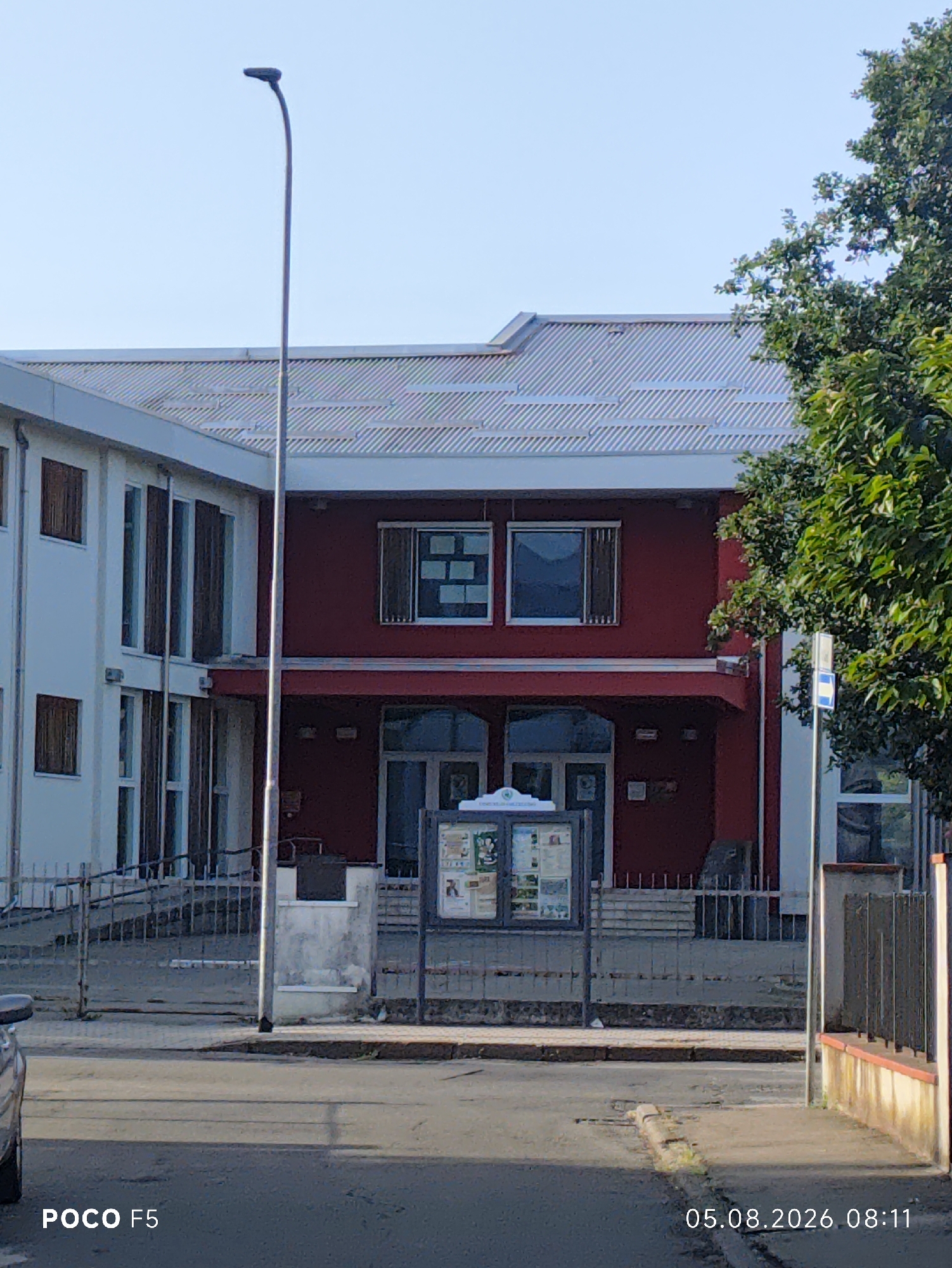 The image shows a street view of a building in Collecchio, Italy. The building appears to be a two-story structure with a red facade on the right and a white facade on the left. There are several windows visible, some with dark shutters. A street lamp stands in front of the building, and a metal fence runs along the sidewalk. A bulletin board with various notices is positioned in front of the red section of the building. A tree with green foliage is visible to the right of the building, and a portion of a car is visible in the lower left corner. The sky is clear and blue. The time stamp on the image reads "05.08.2026 08:11", indicating the photo was taken on August 5, 2026, at 8:11 AM. The "POCO F5" watermark suggests the photo was taken with a Poco F5 smartphone.