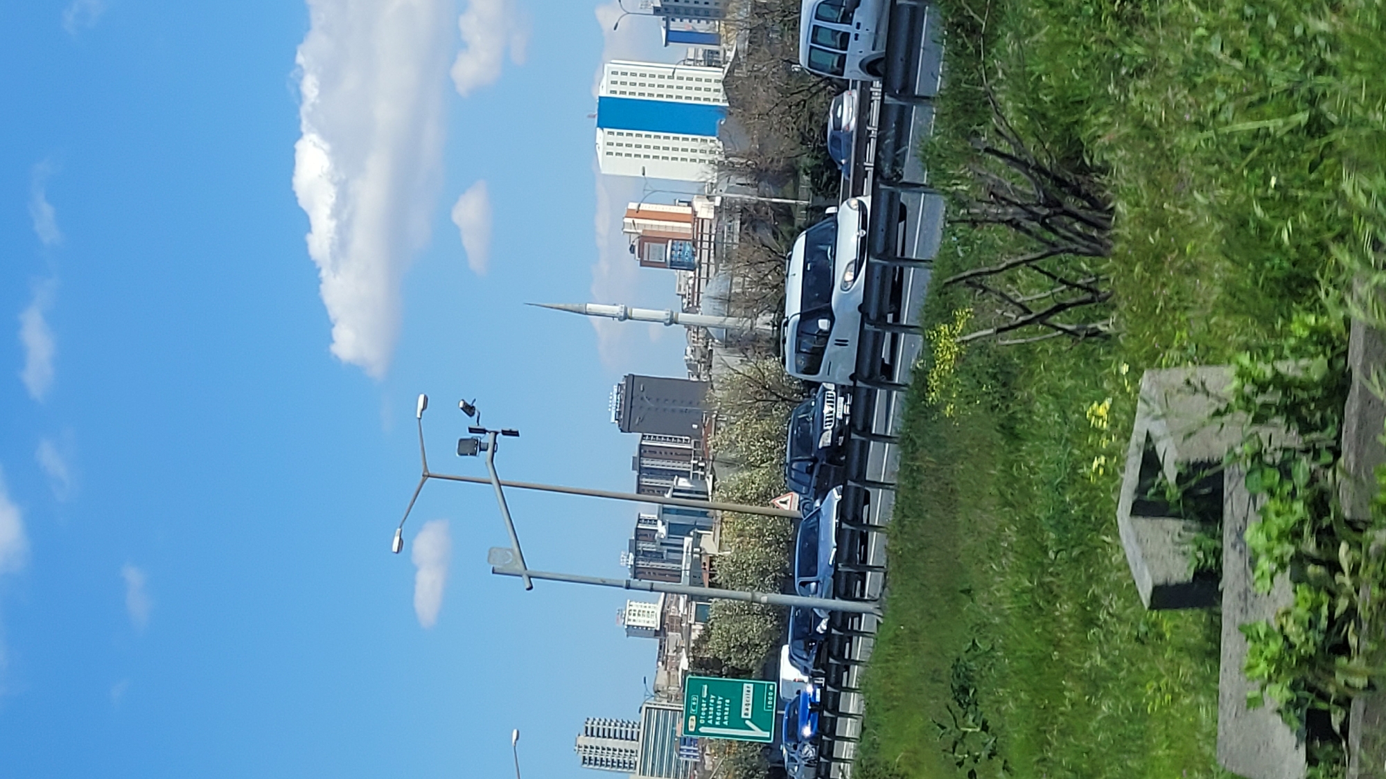 The image captures a view in Bağcılar, Turkey, likely taken from a moving vehicle. It shows an urban environment with a highway running alongside grassy terrain on the right side. Several vehicles are present on the highway, including cars and vans. In the background, multiple buildings are visible, including a mosque with a prominent minaret. Street lamps and road signs are positioned along the highway. The sky is blue with scattered clouds.