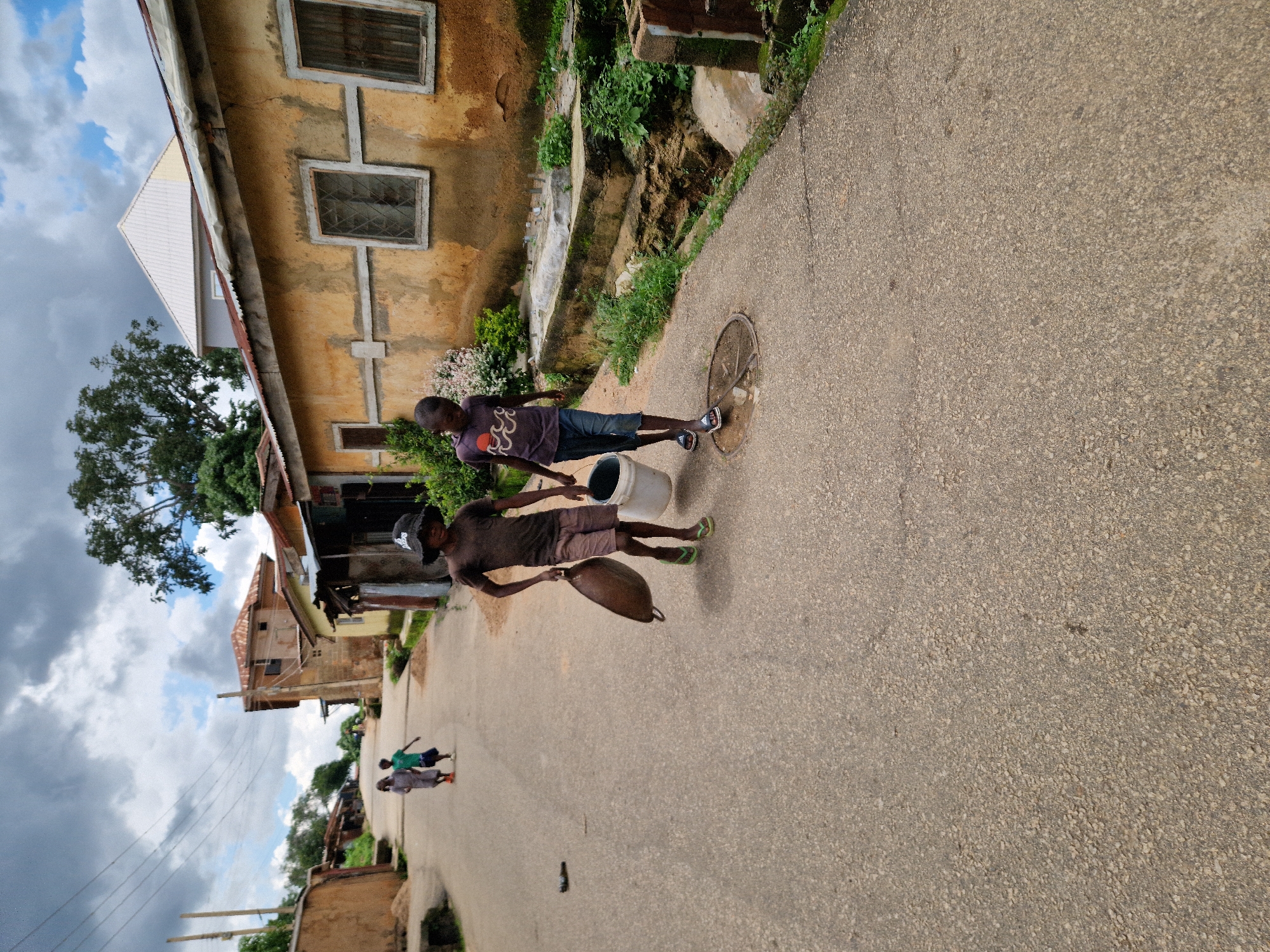 Two young boys are walking down a paved street in Kabong, Nigeria, carrying containers. The boy on the left, wearing a dark shirt, shorts, and green flip-flops, carries a large, brown, pot-like object in his right hand. The boy on the right, wearing a purple t-shirt, blue shorts, and sandals, carries a white bucket in his left hand. They are walking towards the viewer, slightly angled to the right. In the background, other children are visible further up the street. The street is lined with buildings constructed with materials typical of the area, including mud bricks and corrugated metal roofs, some with peeling paint. There is vegetation along the side of the road and a drainage ditch. The sky is cloudy, with patches of blue sky visible. The overall impression is a snapshot of daily life in a community in Nigeria.