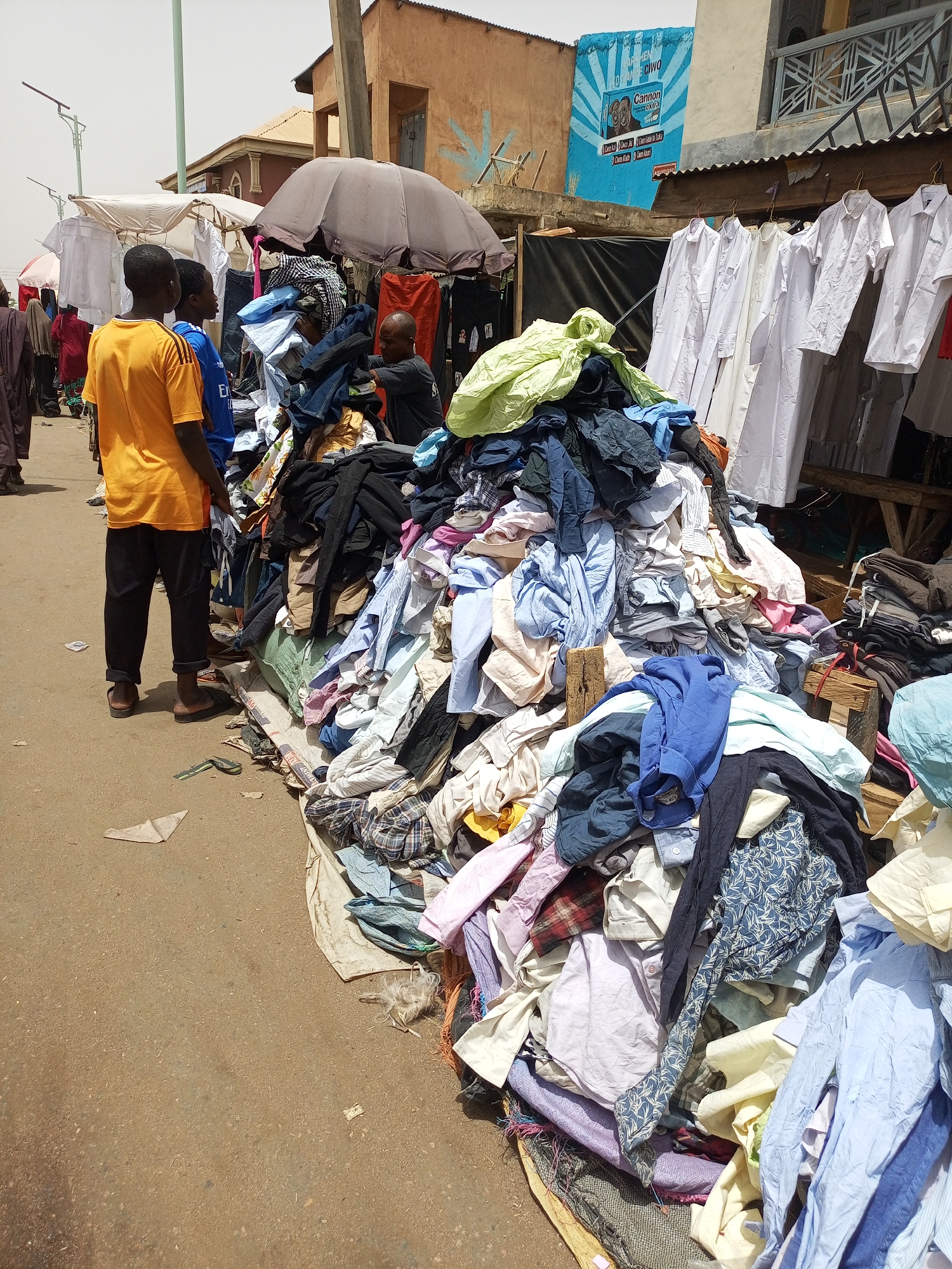 A street scene in Arawa, Nigeria features a large mound of used clothing for sale. Three young men stand to the left, observing the pile. One wears a bright yellow shirt and dark pants. Behind them are other people, stalls with clothing racks displaying white garments, and umbrellas shading the goods. The background shows buildings with faded paint and a faded blue advertisement on one wall. The ground is paved with dirt and gravel.