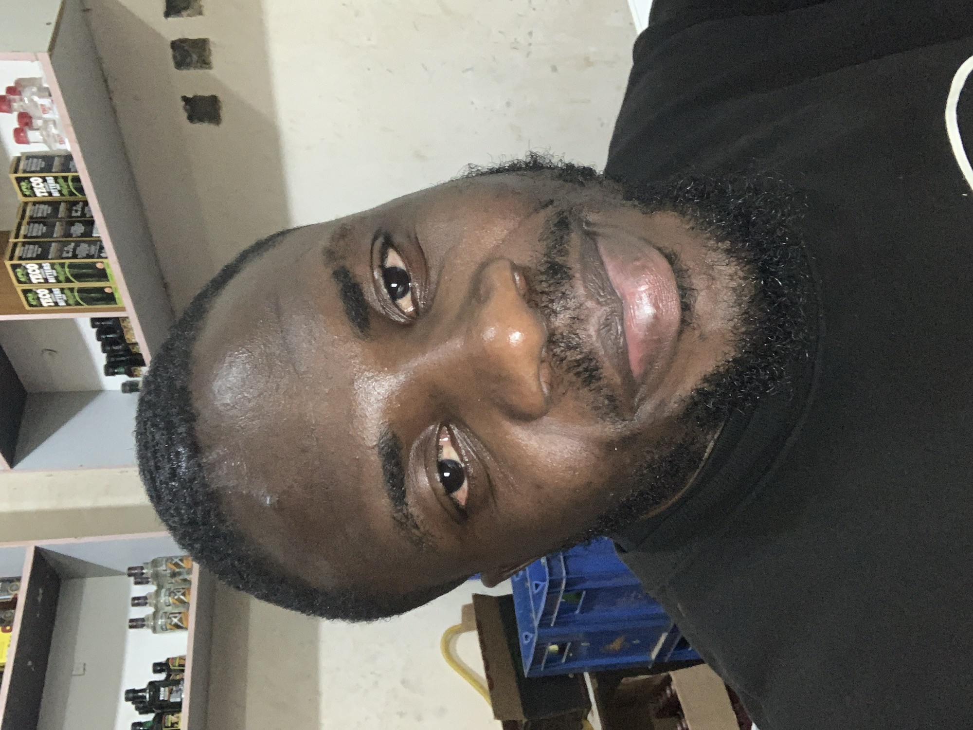 A man with dark skin, short black hair, and a beard smiles at the camera. He is wearing a black t-shirt. The background appears to be a room with shelves displaying various bottles. To the left of the man are two blue crates. The scene is likely indoors, possibly in a home or shop.