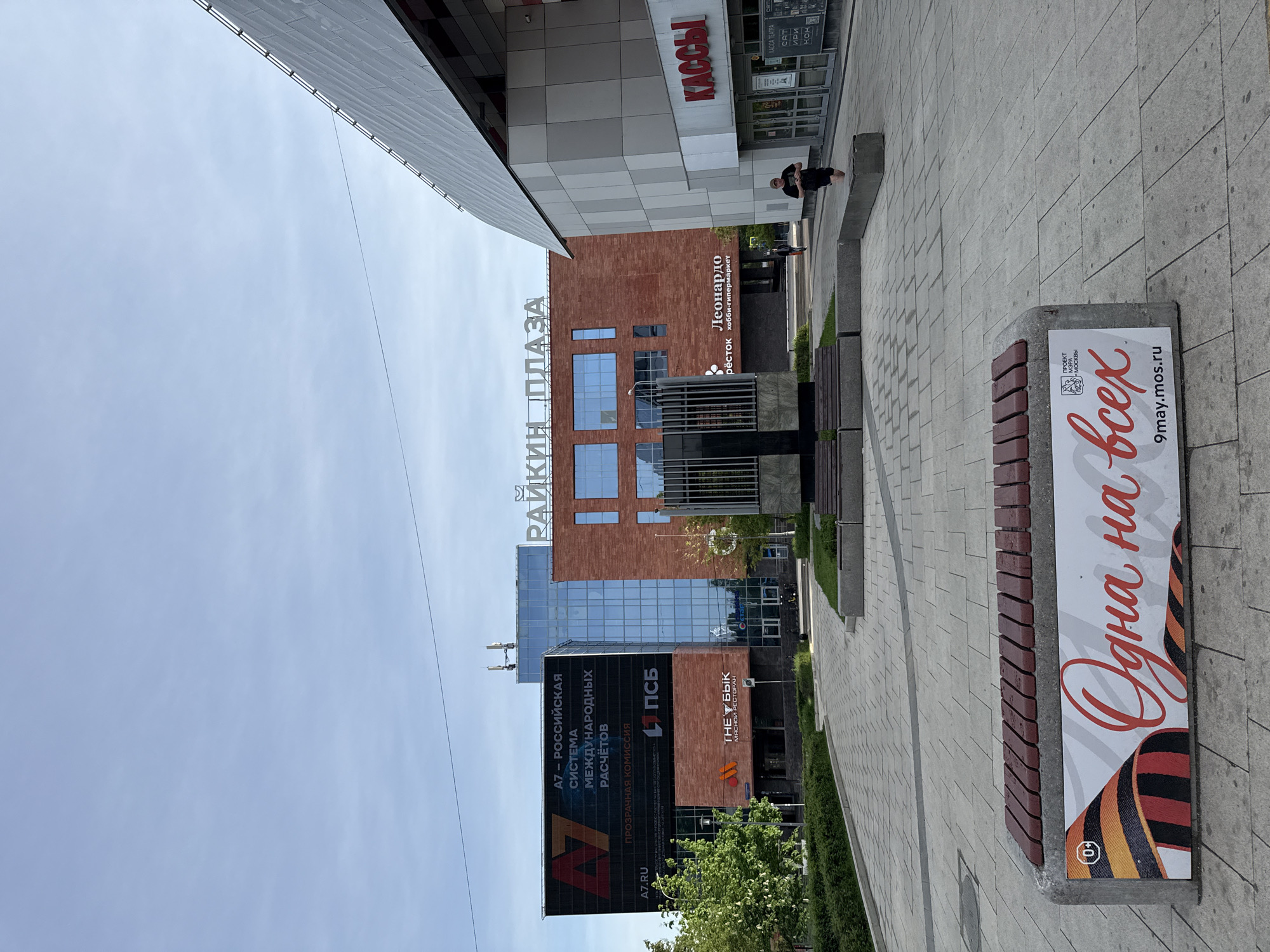 The image shows a street scene in Moscow, Russia. In the background is a multi-story building with a sign that reads "Райкин Плаза" (Raikin Plaza). Other signs on the building include "Леонардо хобби-гипермаркет" (Leonardo hobby hypermarket) and "ПСБ" (PSB). A large advertisement on the building features the text "A7 - РОССИЙСКАЯ СИСТЕМА МЕЖДУНАРОДНЫХ РАСЧЁТОВ" (A7 - RUSSIAN SYSTEM OF INTERNATIONAL PAYMENTS) and "THE БЫК МЯСНОЙ РЕСТОРАН" (THE BULL MEAT RESTAURANT). The image also shows a bench with a sign that reads "Одна на всех" (One for all) and "9may.mos.ru". In the upper right, there is also a sign with "КАССЫ" (Boxes) on it. In the distance, there is a person sitting. The environment appears to be a pedestrian area.