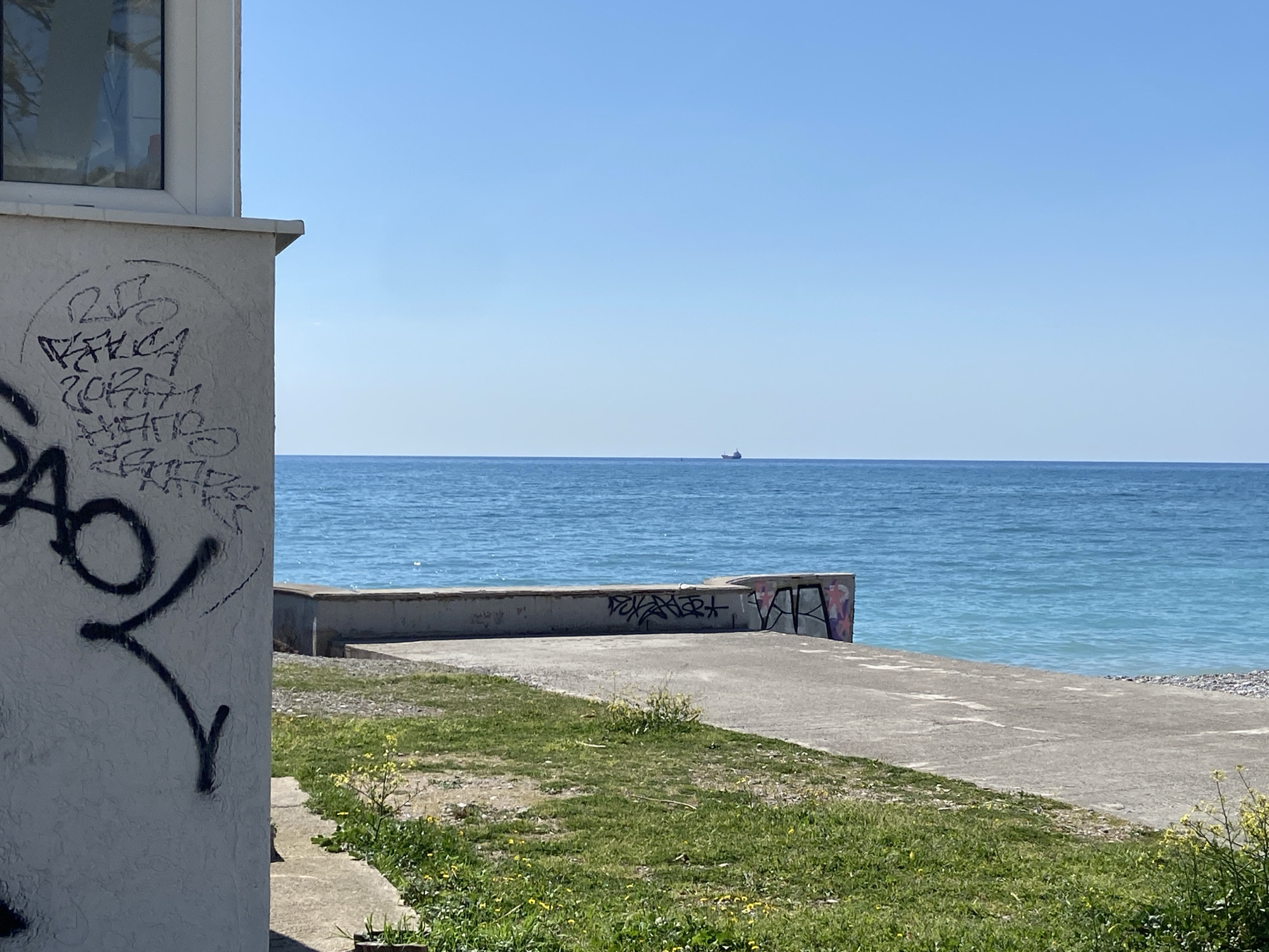 The image shows a seaside scene in Bar, Montenegro. In the foreground is a white building with graffiti on its walls. Next to it is a concrete path with patches of grass growing through it. Beyond the path is a low concrete wall, also covered in graffiti. The Adriatic Sea stretches out to the horizon, with a single ship visible in the distance. The sky is clear and blue.