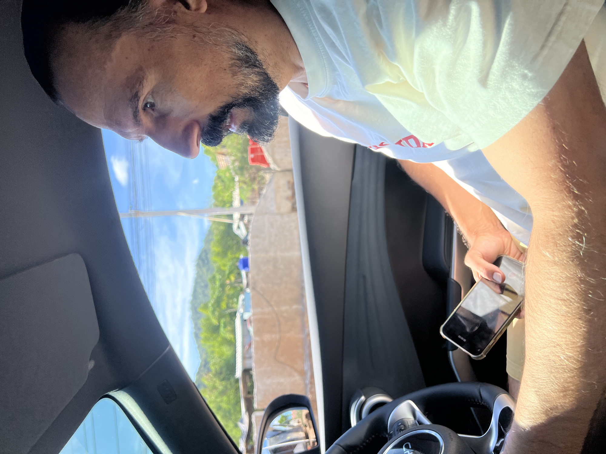 A man with a dark beard and mustache is sitting in the driver's seat of a car, holding a cell phone in his right hand. He is wearing a white t-shirt. The interior of the car is visible, including the steering wheel and dashboard. Outside the car window, there is a view of a neighborhood with buildings, trees, and power lines. The location is identified as the city of Unknown, Unknown.