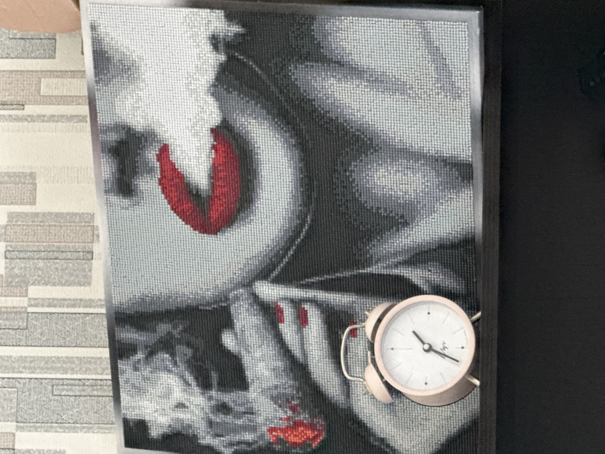 A pixelated, framed image depicting a person with red lips and red nails is placed on a surface with a light brown and gray patterned covering. An alarm clock with a white face and pink trim is positioned in front of the picture, obscuring the lower right area. The alarm clock reads approximately 7:12. The background to the right of the image is black.