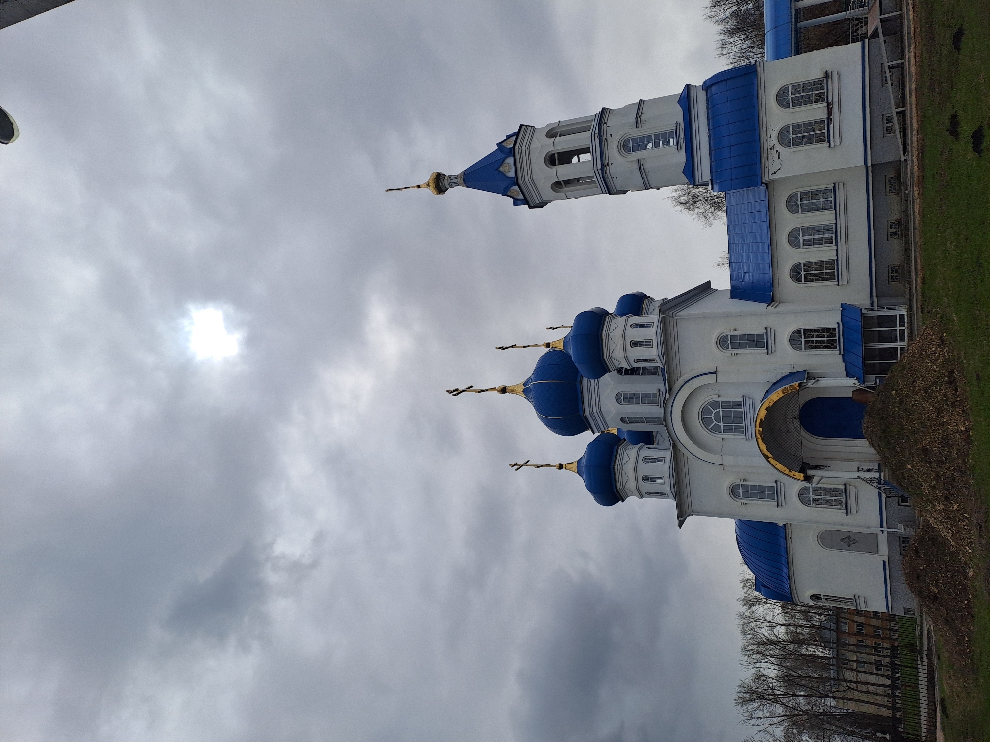 This image captures a prominent Orthodox church in Yalga, Russia, under an overcast sky. The church features striking white walls contrasted with vibrant blue roofs and multiple blue onion-shaped domes, each crowned with a golden Orthodox cross. A taller bell tower with a similar blue spire and golden cross stands to the right of the main building structure.
The sky is heavily clouded, presenting various shades of grey, with a bright, diffused spot of light visible through the dense clouds on the upper left, suggesting the sun attempting to break through. The overall lighting is soft and somewhat dim.
The ground in the foreground and to the right shows areas of dry grass and scattered fallen leaves, indicative of late autumn or early spring. Bare trees are visible behind a dark fence line on the left side of the church, further supporting the seasonal observation. There are no people visible, and no discernible activity is taking place. The image is a static architectural photograph. No visible text is present on the building or in the surroundings.