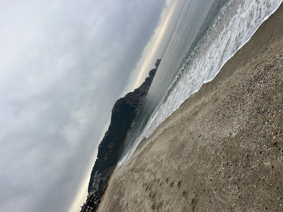 The image displays an outdoor scene of a beach and coastline, identified as Kleopatra Beach in Alanya, Turkey, under an overcast sky. The foreground consists of a wide expanse of sand mixed with small pebbles, exhibiting multiple human footprints. The ocean occupies the midground and right, with small, white-crested waves breaking along the shoreline against the sand. The water surface is a muted grayish-blue. In the background, a large, green, tree-covered hill rises on the left, displaying several multi-story buildings with dark roofs and light facades. This hill transitions into rugged, dark rocky cliffs that extend into the sea towards the right, forming a peninsula. The sky above is uniformly covered by a dense, light-grey cloud layer, indicating overcast weather conditions. No individuals are visible within the frame.