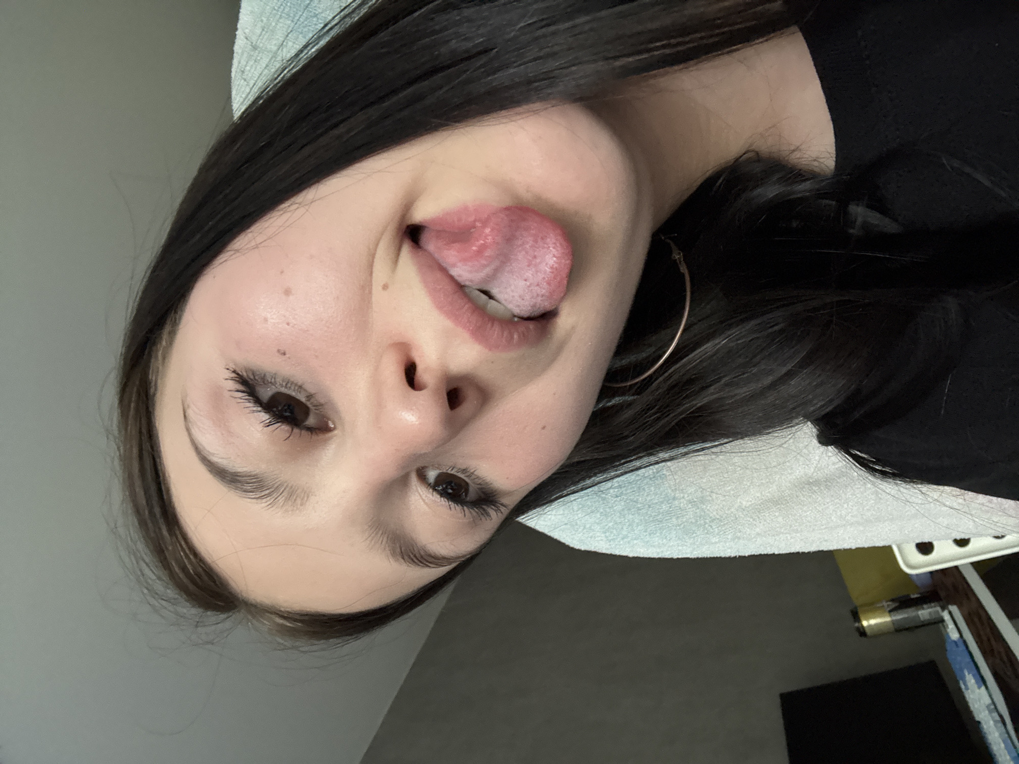 A young woman with dark hair and light skin is making a silly face, sticking her tongue out. She is wearing a black shirt and gold hoop earrings. A light blue towel is draped behind her head. The background is blurry but appears to be a room with neutral gray walls. The location is not identifiable as the city of Chaikovsky, Russia based on the provided image.