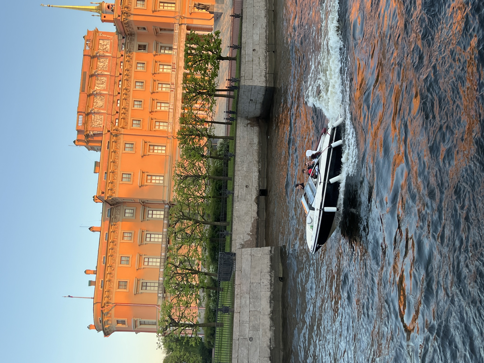 The image captures a scene in Saint Petersburg, Russia, showcasing the Mikhailovsky Castle (also known as the Engineers' Castle) along a canal. The castle's façade is visible on the left, displaying its characteristic orange hue and ornate architectural details. A row of trees lines the embankment in front of the castle, separated from the canal by a stone wall and a wrought iron fence.
To the right, a motorboat cuts through the water, creating a visible wake. Figures are visible within the boat, suggesting people are aboard. The water reflects the colors of the sky and buildings, with shades of orange and gray. The overall lighting suggests the scene is captured during the late afternoon or early evening, as the sun casts long shadows and illuminates the castle with a warm glow.