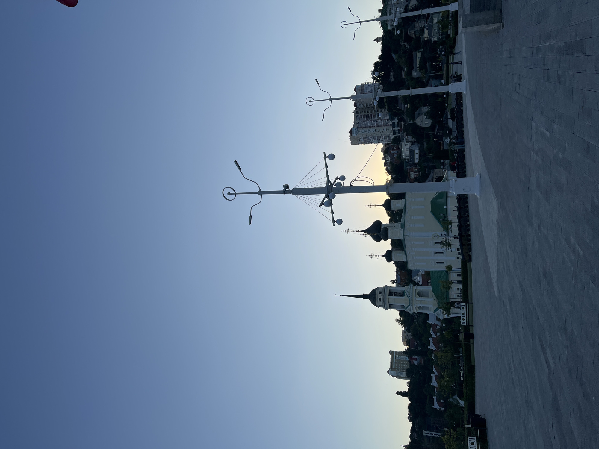 The image captures a wide-angle view of a public square in front of a hillside lined with buildings, including churches and residential structures. The sky is clear and blue, suggesting it is either early morning or late evening. Several modern streetlights with multiple lamps are prominently featured in the foreground, their silhouettes stark against the sky. In the mid-ground, two domed churches with crosses atop are visible, one taller with a bell tower. Beyond the churches, a multi-story residential building stands out on the hillside, with smaller houses and trees scattered across the slope. The ground in the foreground is paved with large, grey rectangular tiles. Along the edge of the paved area, a line of old cannons is displayed, suggesting a historical or memorial context. A few indistinct figures of people can be seen in the distance, walking or standing. The overall scene conveys a sense of urban tranquility and architectural interest. This scene is located in Novaya Usman, Russia.