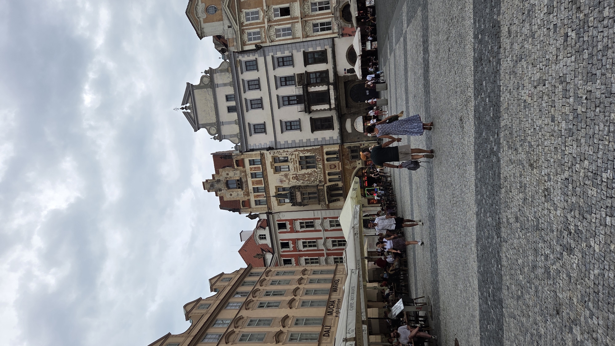 The image shows a view of Old Town Square in Prague, Czechia, on an overcast day. In the foreground, a cobblestone plaza is visible, with two couples walking away from the camera. The couple on the right is holding hands, with the woman wearing a blue and white floral dress and the man in a dark t-shirt and khaki shorts. The couple on the left consists of a man in a white shirt and shorts and a woman in a white top and a dark skirt.
Further back, several buildings with distinctive architecture line the square. To the left, a large building with light-colored facade and multiple windows is prominent. Next to it, a red and white building with several windows stands out. The central building features intricate facade decorations and a stepped gable. To the right, another building with a light-colored facade and multiple windows is visible.
Several outdoor cafes with white umbrellas and seating are set up in front of the buildings, with many people seated at the tables. Some signs are visible above the cafes, including "DALI MUCHA WARHOL exhibition" and "CAFFE-RISTORANTE". The sky is filled with clouds, creating a diffused light across the scene.