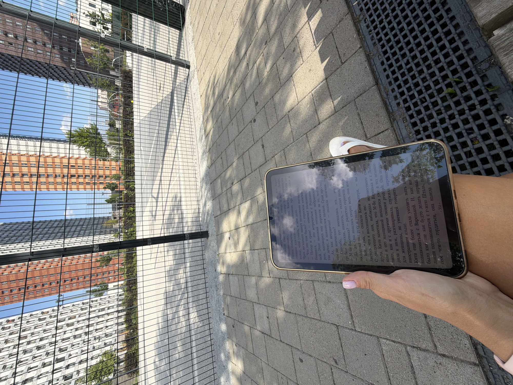 The image shows a person's hand holding a tablet device displaying a Russian language text. The tablet is resting on the person's lap, with their foot wearing a white sandal visible in the lower right corner. The background features a paved area with a metal grate, and beyond that, a fence and several modern residential buildings. The buildings are tall, with many windows and visible architectural details, suggesting an urban environment. The sky is blue and clear. The overall impression is of a person reading outdoors in a contemporary urban setting, likely a park or a recreational area within a residential complex. The text on the tablet is largely illegible due to the angle and resolution, but it appears to be a novel or story. The scene suggests a moment of leisure and relaxation.