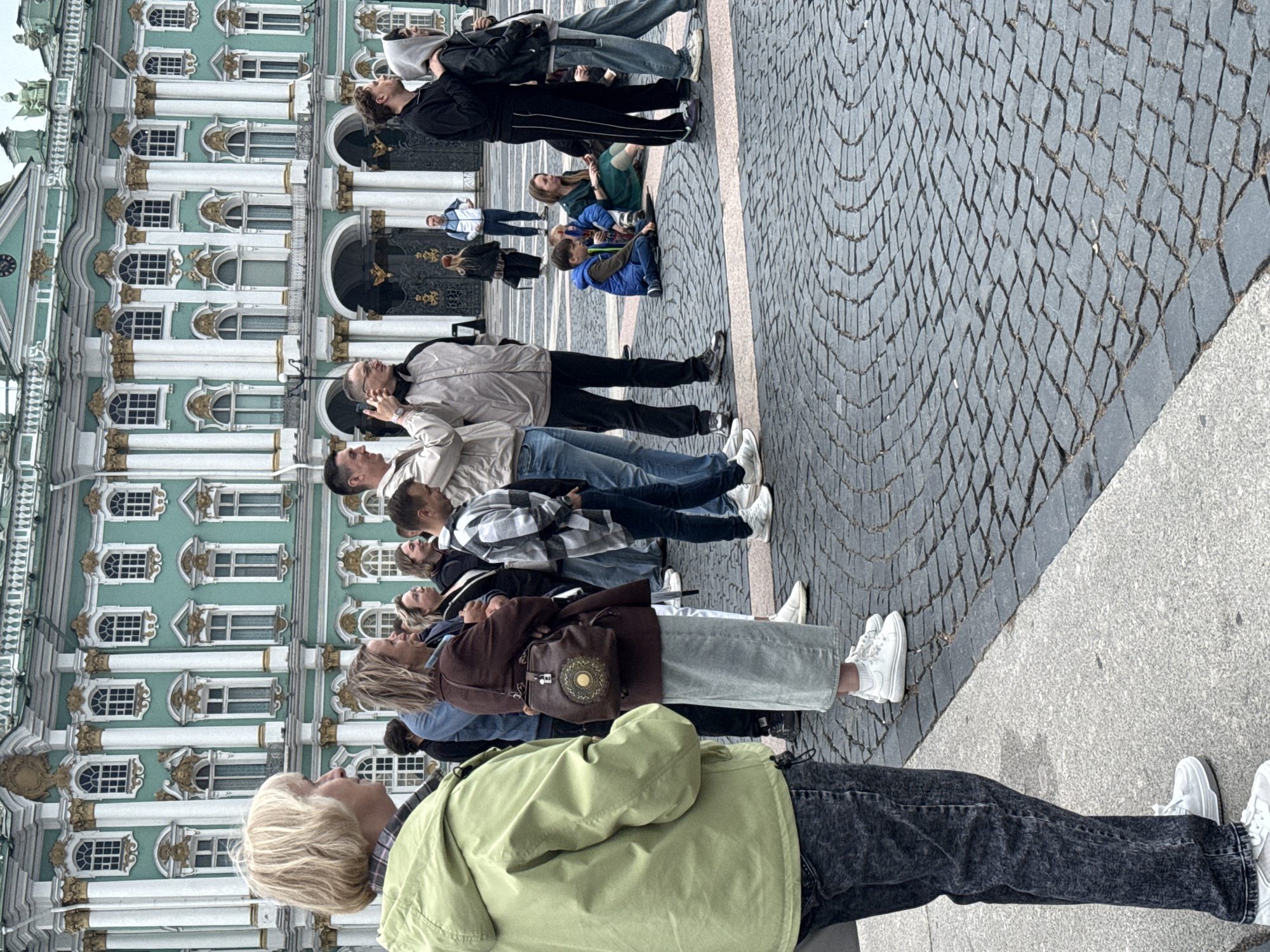 A group of people are standing and sitting on a cobblestone plaza in front of a large, ornate green and white building with gold accents. The building appears to be a grand palace or museum, characterized by its classical architecture, numerous windows, and columns. The plaza is paved with dark grey cobblestones arranged in a circular pattern in some areas.
In the foreground, a woman in a light green hooded jacket and dark, distressed jeans stands facing to the right, looking towards the building. Beside her and slightly behind, a woman in a brown jacket and a long, light blue skirt is also facing right, with her arms crossed. Further back and to the right, a group of people are visible, including men and women standing and two children sitting on the ground. Some individuals are looking towards the building, while others appear to be engaged in conversation or taking in the surroundings. The overall atmosphere suggests a tourist or public gathering, likely in a prominent city location. Based on the architectural style of the building, it is highly probable that the location is the Hermitage Museum (Winter Palace) in Saint Petersburg, Russia.