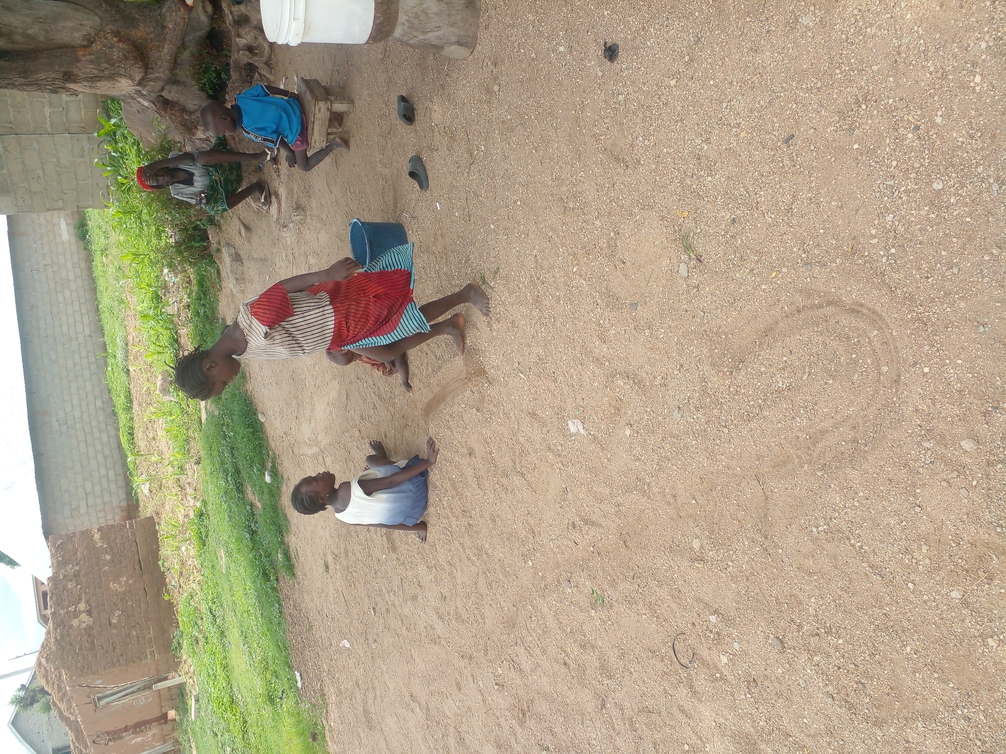 In an outdoor, dusty environment with some green vegetation and partially constructed buildings in the background, four individuals are present. In the foreground, two young children are sitting on the ground. One child, on the left, is wearing a white and blue dress. The other child, further to the right, is standing and wearing a striped top and a red and blue skirt. A blue bucket is near the standing child. To the right of the standing child, another child, a boy wearing a blue shirt and red shorts, is sitting on a small wooden stool. Behind the boy, an adult woman with a red headscarf is sitting, with her hand on her head. In the background, there are structures made of mud bricks and concrete blocks, and a large tree is visible on the right. Several pairs of sandals are on the ground near the adults and the stool. A large white bucket is situated to the far right. The ground is covered in dirt and small pebbles.