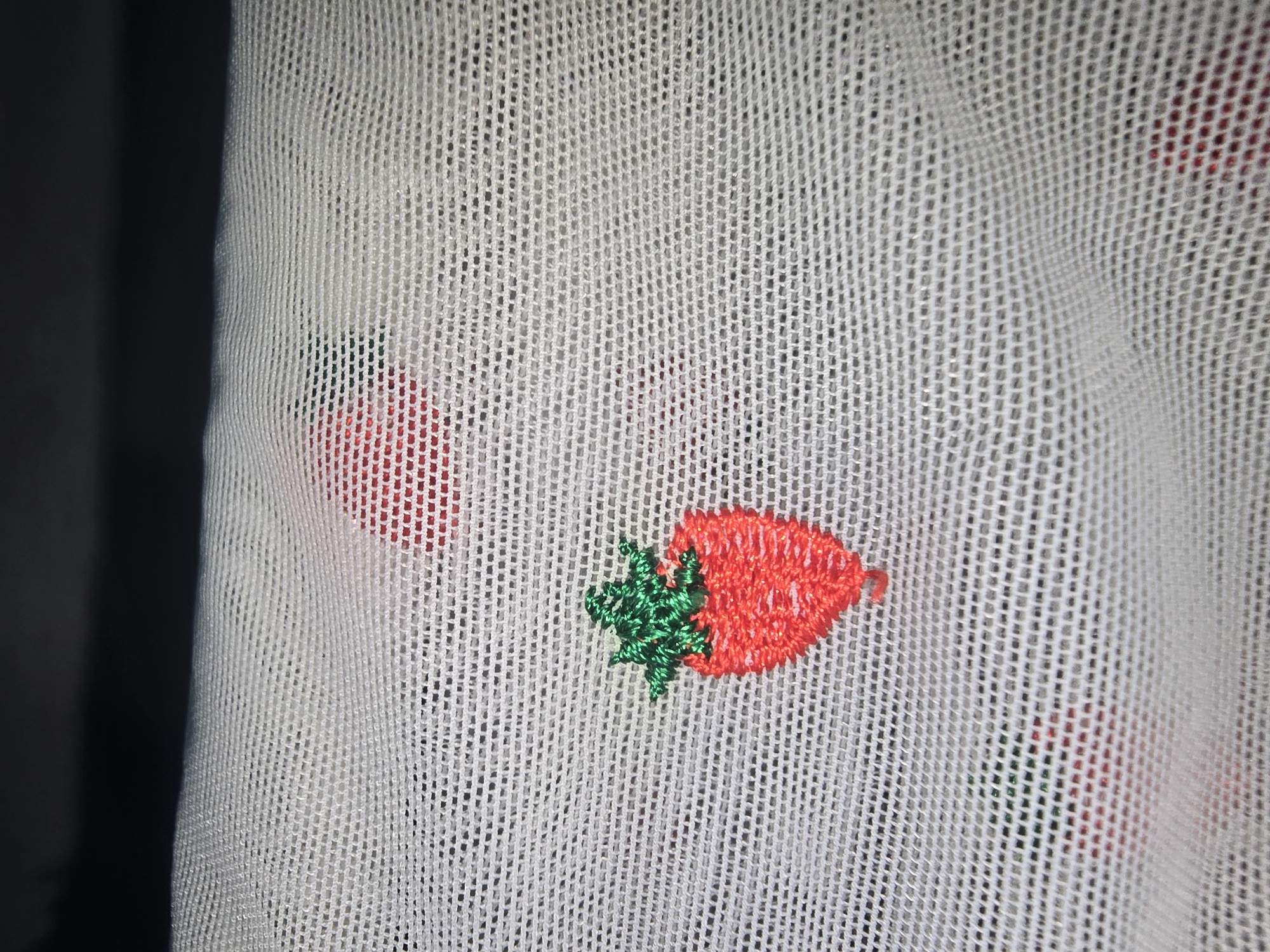 The image shows a close-up view of white mesh fabric with embroidered strawberries. The strawberries are small, with red bodies and green stems and leaves. Some of the strawberries are partially visible, suggesting a pattern. The fabric appears to be a delicate material, possibly used for clothing or home decor. There is no indication of the image's location in Ulan-Ude, Russia, or any contextual information about people, actions, or the environment.