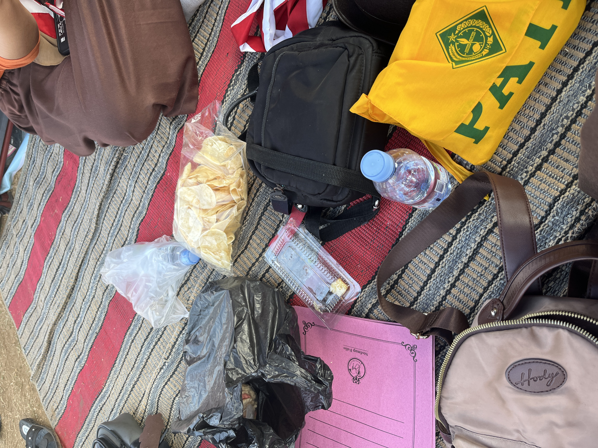 This is a close-up, eye-level shot of a group of items laid out on a striped mat on the ground. The mat has alternating stripes of red, beige, and gray. The items suggest a casual outdoor gathering or picnic.
In the foreground, a brown fabric bag with a zipper is visible, its strap extending upwards and to the right. To the right of this bag is a bright yellow fabric bag with green lettering that appears to spell out "PAI" along with a circular green and yellow emblem featuring text. A clear plastic water bottle with a blue cap is positioned to the left of the yellow bag.
Further into the image, a black shoulder bag with multiple compartments and straps is prominent. Next to it, a pink folder or notebook lies flat. To the left of the pink folder, a black plastic trash bag is partially opened, revealing some contents. Above the trash bag, a clear plastic bag filled with light-colored potato chips is visible.
In the upper right portion of the image, a person's legs are visible, wearing loose brown trousers. Their arms, in an orange top, are reaching towards something out of frame. A small black electronic device, possibly a remote, is also visible near the person's hand.
The scene appears to be an outdoor setting, likely a park or gathering space, given the mat and picnic-like arrangement of items. The environment is natural, with a hint of dry earth visible around the mat, suggesting a dry climate. The location is Gembong, Indonesia, and the objects indicate a possible recreational activity or a break during an event.