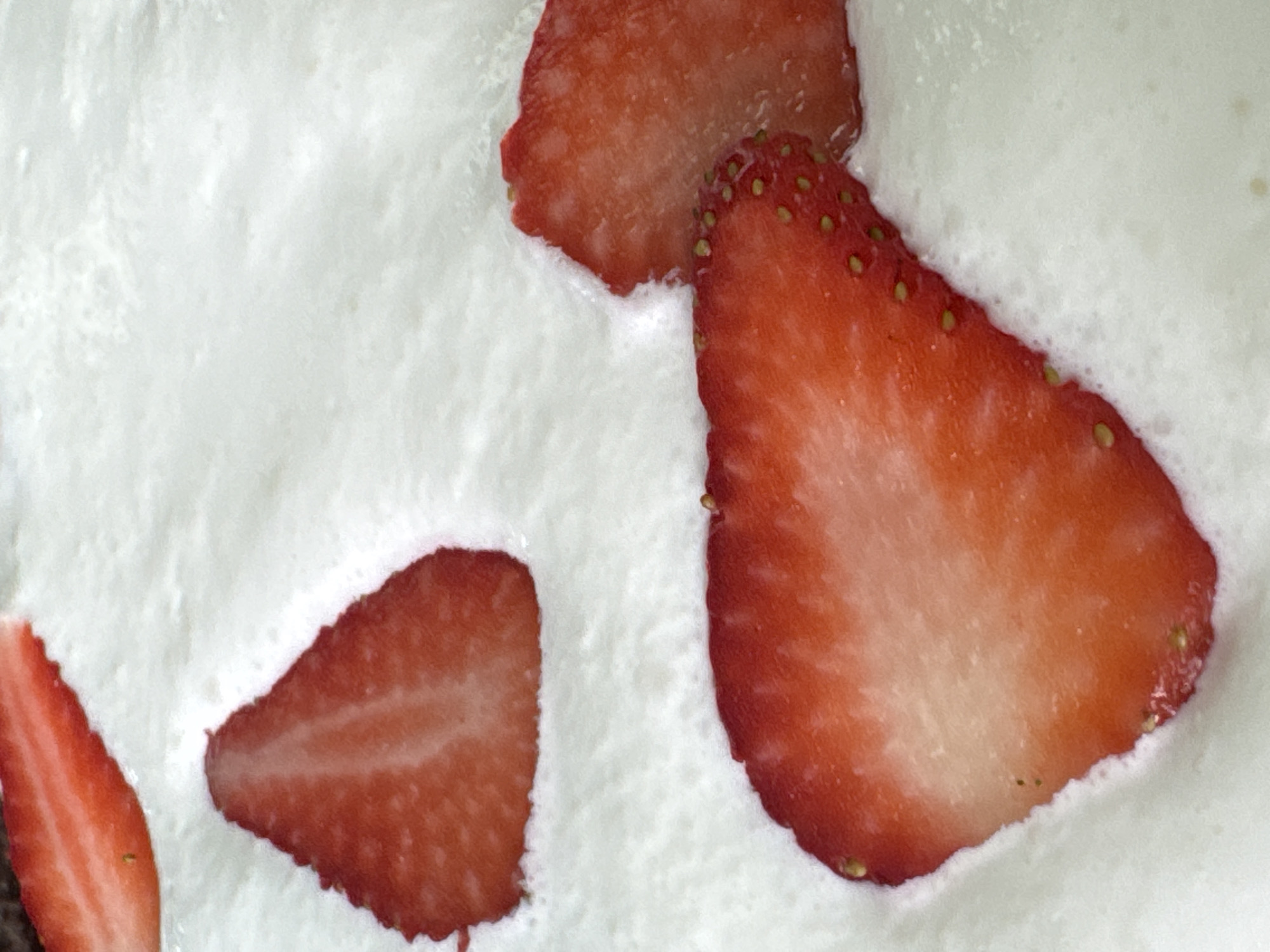 A close-up photograph shows sliced strawberries on top of a white, textured surface resembling whipped cream or frosting. Four strawberry slices are visible, each cut in half to reveal the interior. The strawberries range in color from light red at the core to a darker red at the edges, with visible seeds on the outer surface. The creamy white surface has a bubbly, irregular texture. The setting is likely indoors, but the specific location in San Bernardino Tlaxcalancingo, Mexico is not discernible from the image alone.