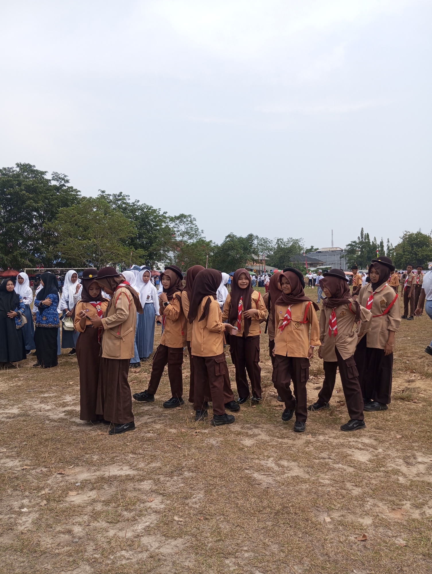 This is a full-shot, eye-level photograph taken outdoors on a cloudy day. The scene depicts a group of young people, likely students, dressed in brown scout uniforms with red and white neckerchiefs. They are standing in a dry, grassy field with sparse vegetation. In the background, there are trees, buildings, and a few other people in the distance. Some of the students are looking towards the camera, while others are engaged in conversation or looking elsewhere. The overall impression is of an outdoor gathering or event. While the location is specified as Pematang Reba, Indonesia, there are no specific landmarks visible in the image that would allow for precise identification of a particular spot within the city.