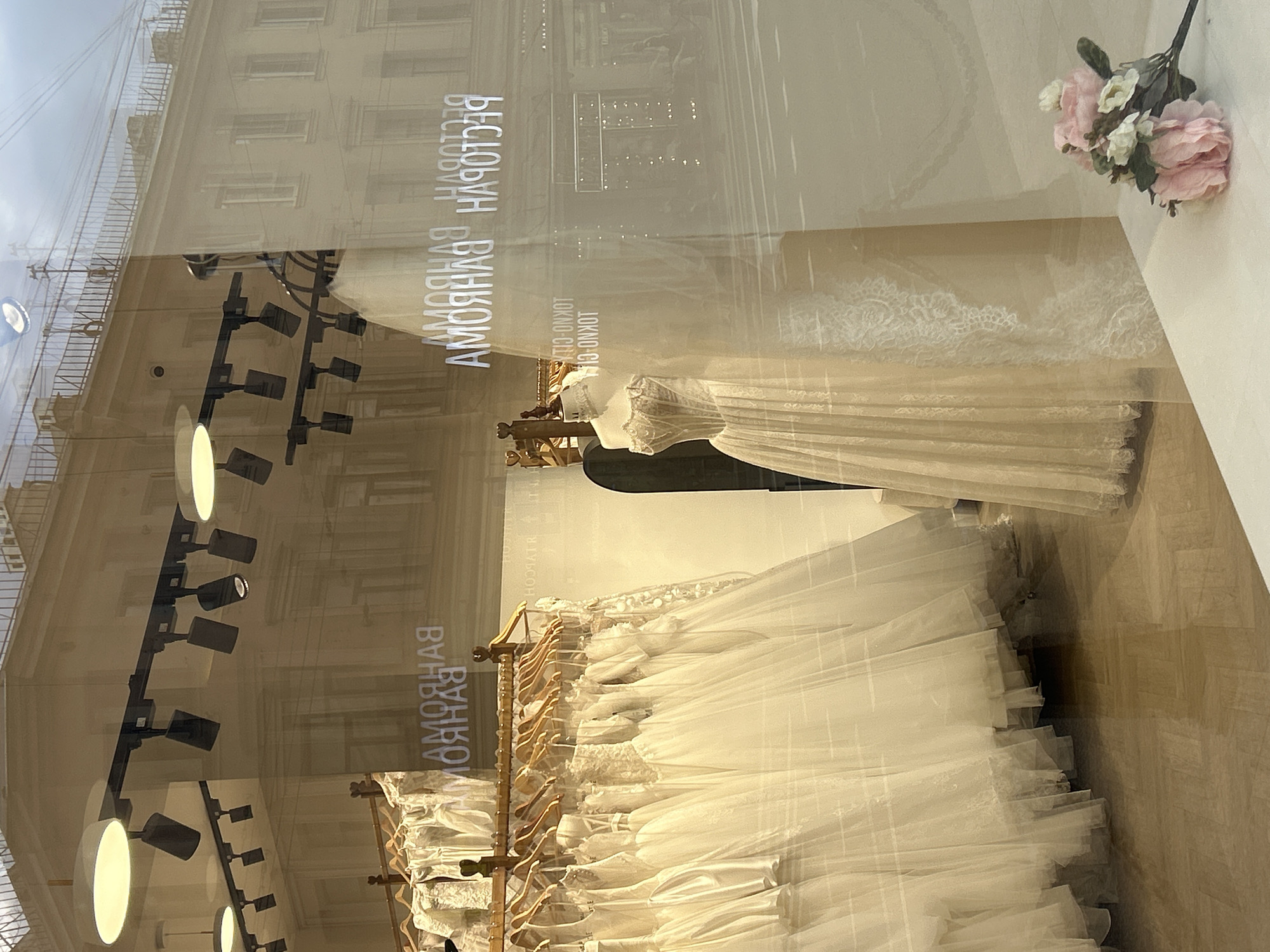 The image shows the interior of a bridal shop, viewed through a large window with reflections. Inside, numerous white wedding dresses are hanging on a wooden rack. One dress is displayed on a mannequin, a formal gown with intricate lace detailing. In the foreground, a bouquet of pink and white artificial flowers rests on a white surface. The reflections on the window show buildings and a cloudy sky, suggesting the shop is located in a city. The text "РЕСТОРАН" and "АМОЯЯЯ" is visible in reverse through the glass, possibly indicating the name of the shop or a nearby establishment. The floor appears to be polished wood. The lighting within the shop comes from track lighting fixtures mounted on the ceiling.