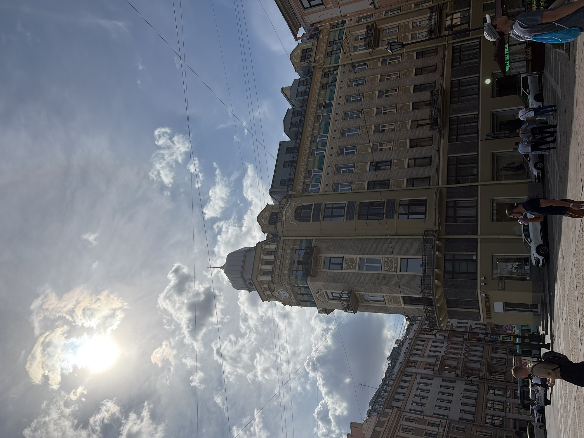 The image depicts a street scene in Saint Petersburg, Russia, with tall, ornate buildings lining a pedestrian walkway. The perspective is from ground level, looking upwards towards the sky, which is filled with dramatic, sun-drenched clouds. The sun is partially obscured by clouds, suggesting it's either morning or late afternoon. The architecture is detailed, with multiple stories, balconies, and decorative elements on the building facades. Power lines crisscross the sky, indicating an urban environment.
Several people are visible on the sidewalk, walking and standing. One person in the foreground is seen from behind, wearing a dark shirt and backpack. Further down the street, a small group of people are gathered, and another individual stands near a parked white car. The overall atmosphere is that of a bright, sunny day in a European city.
Visible text includes "II Collection" on a storefront. Another sign in Cyrillic appears above a doorway, though it is partially obscured and difficult to read clearly. The image captures a moment of everyday life in a city with rich architectural heritage.