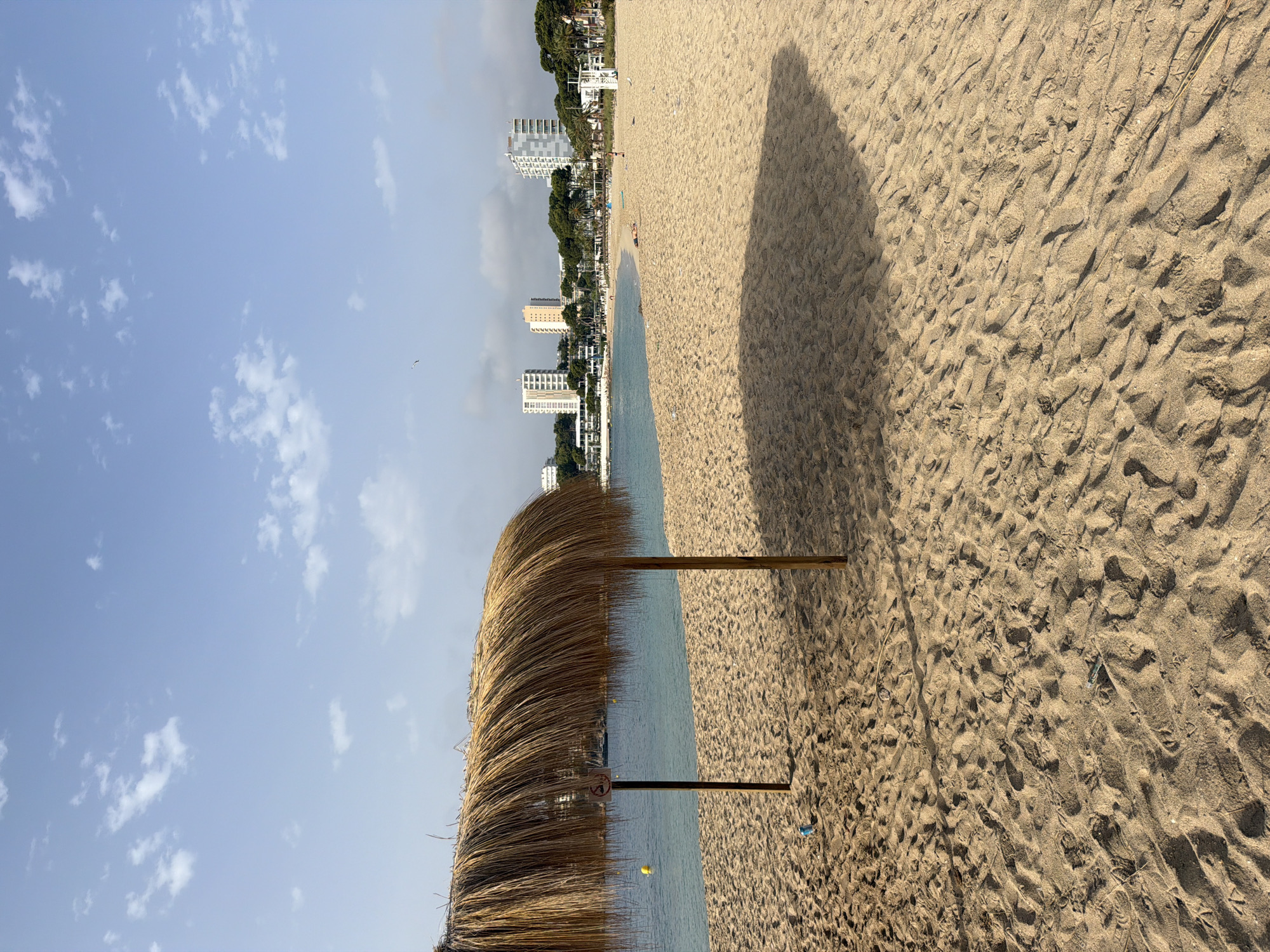 The image depicts a sunlit beach in Magaluf, Spain. In the foreground, a thatched umbrella provides shade over a sandy expanse. The sand is marked with footprints and has a rough texture. To the left of the umbrella, a yellow buoy floats in the calm, turquoise water. In the background, several white buildings, including hotels and apartment complexes, are visible, backed by lush green trees. A lifeguard tower stands on the right side of the beach. A few people are scattered across the sand, with one person lying down near the water. The sky is a clear blue with scattered white clouds.