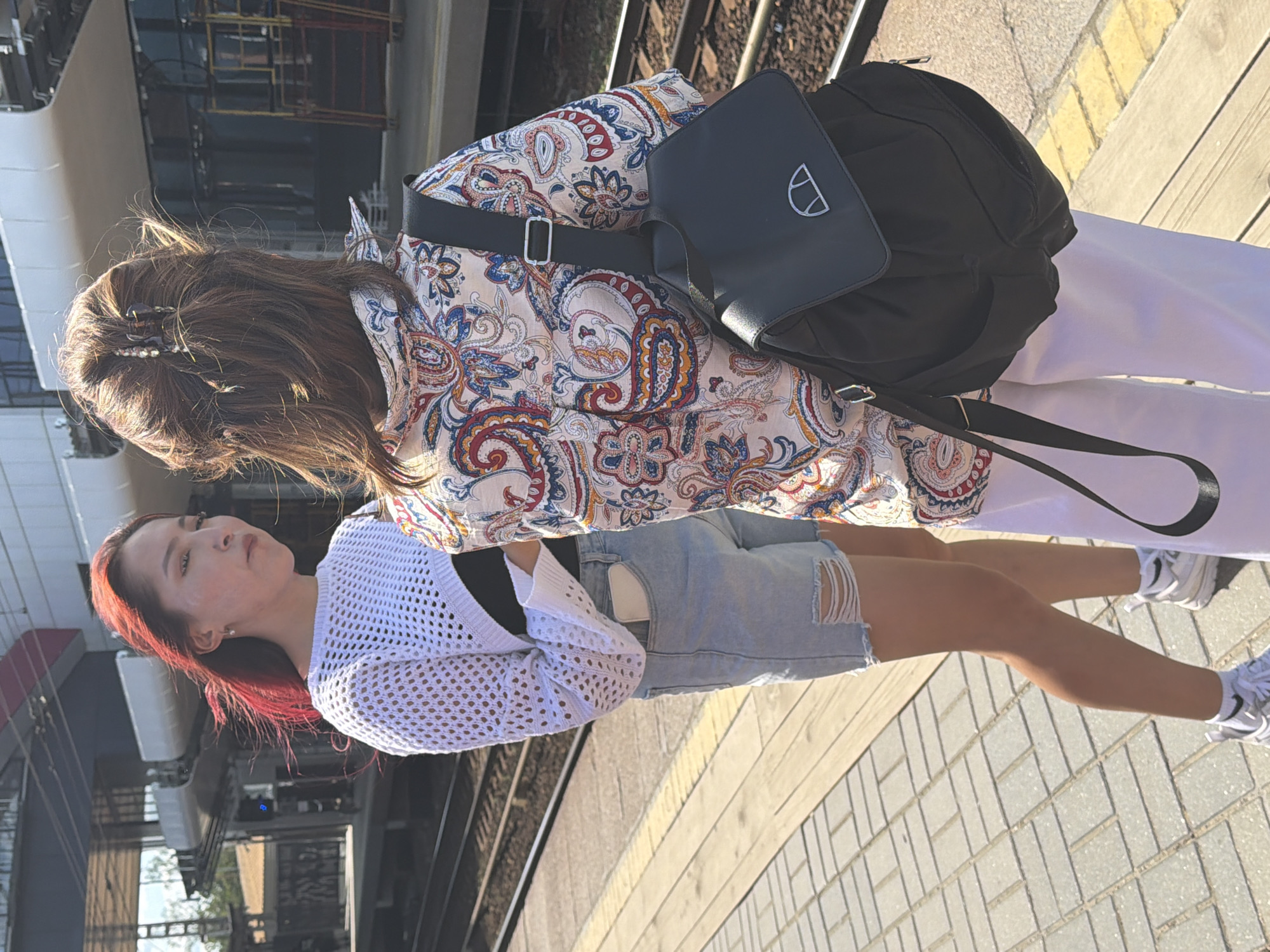 Two young women stand on a train platform. The woman on the left has red hair and wears a white crocheted sweater over a black top and ripped denim shorts. The woman on the right has brown hair and wears a patterned button-down shirt, white pants, and a black backpack. They are both looking away from the camera, seemingly waiting for a train. The platform is made of wood and tile, and train tracks are visible in the background, along with a train station structure. The lighting suggests it is daytime.