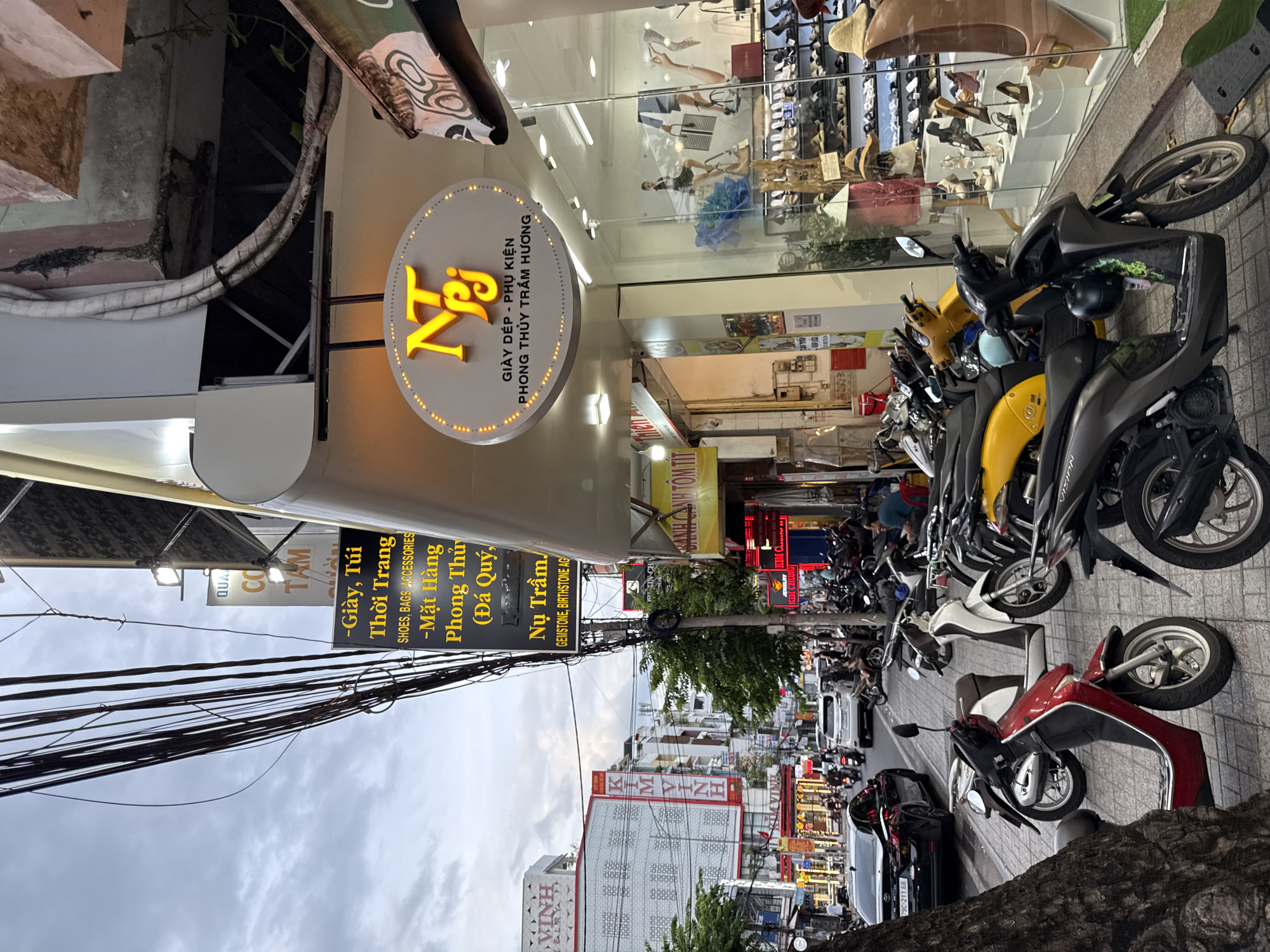 The image captures a street scene in Nha Trang, Vietnam, under an overcast sky. A busy sidewalk is lined with parked motorcycles, and a few cars are visible on the road. Several shops are open, with signs in Vietnamese and English advertising products such as shoes, bags, accessories, and gemstones. The shop fronts are decorated with displays of merchandise, including shoes and handbags. A prominent sign for "NTj" features a yellow logo and text in Vietnamese, indicating a shop selling shoes, accessories, and gemstones. Overhead, a tangle of electrical wires crisscrosses the sky. People are sparsely present on the sidewalk, with some individuals walking or sitting. The overall impression is of a commercial street with active retail businesses.