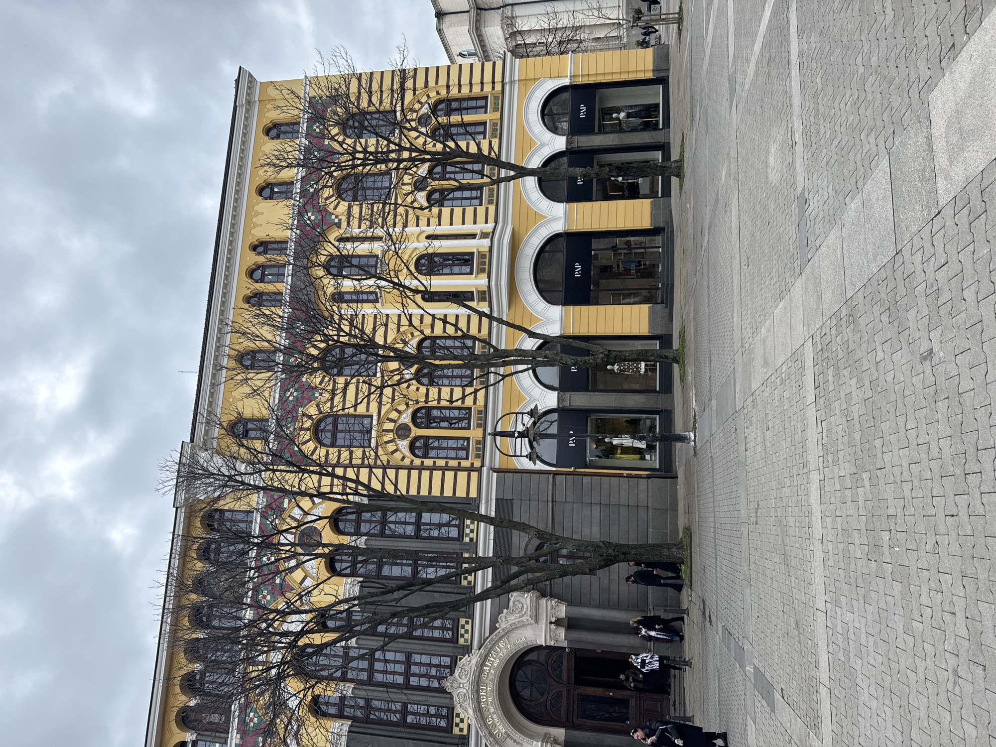 The image shows a building in Sofia, Bulgaria. The building has a yellow facade with arched windows, and some of the ground-level spaces are occupied by shops. Bare trees stand in front of the building, obscuring some of the facade. People are walking on the cobblestone pavement in front of the building. The sky is overcast.