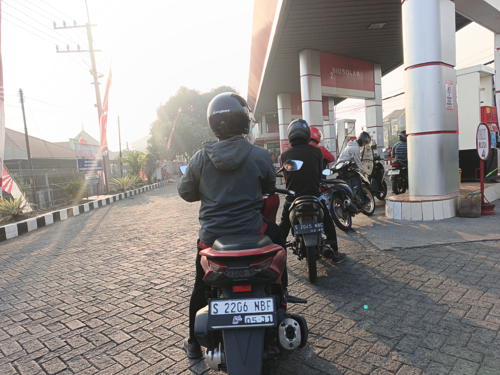 In Pacing, Indonesia, at a gas station, several people on motorcycles are gathered. The sun is low in the sky, casting long shadows across the cobblestone pavement. The gas station features red and white structures, with signs indicating "BIOSOLAR" and pump numbers. One motorcyclist in the foreground, facing away from the camera, wears a black helmet and a grey jacket. Another motorcyclist behind them wears a black helmet and a dark top. A third motorcyclist in the background wears a red helmet. Several motorcycles are parked, including one with a license plate visible that reads "S 2045 NBC". Trees and buildings are visible in the background, suggesting an urban or suburban environment.