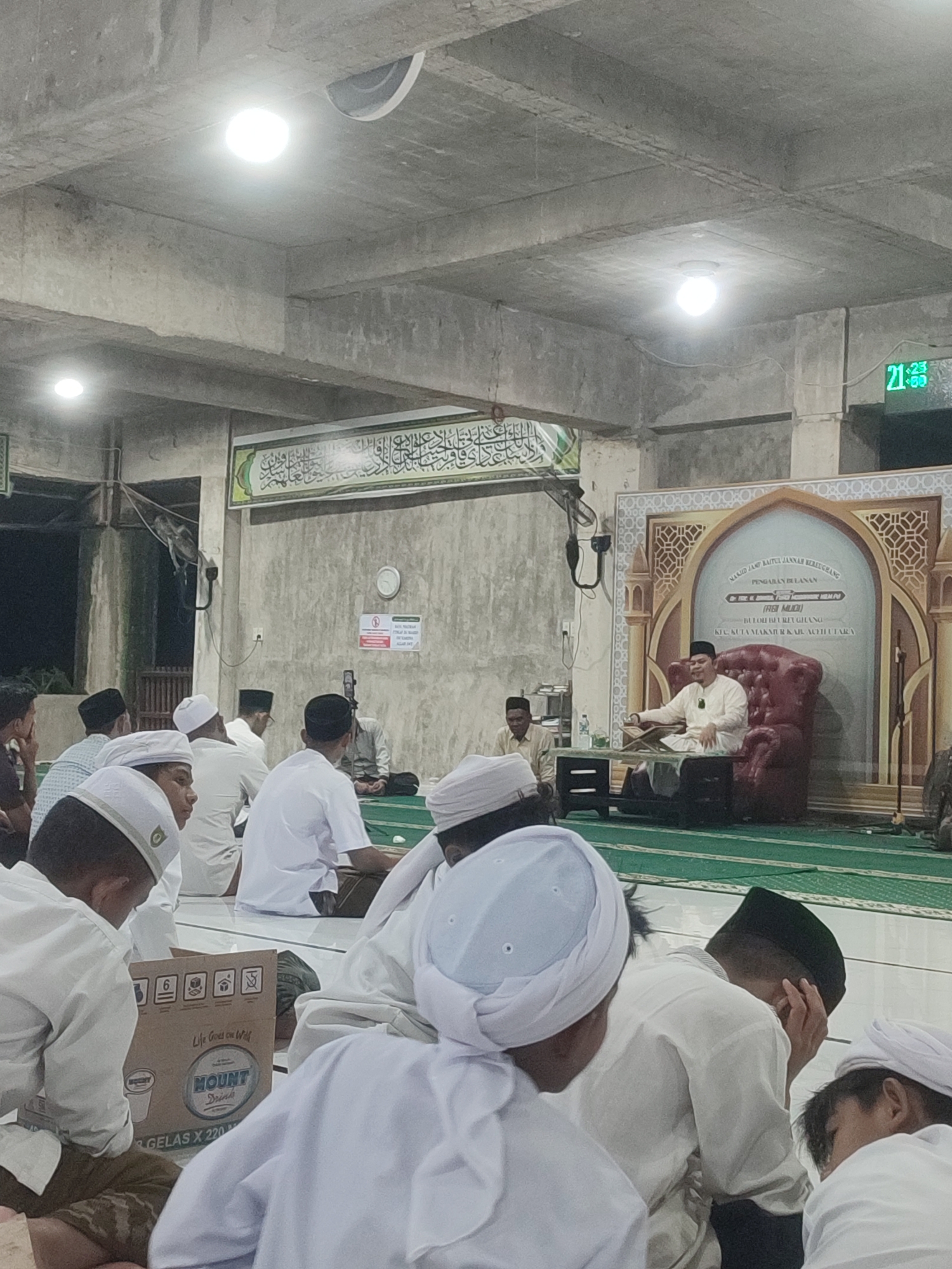 The image shows a religious gathering inside a mosque. Many men and boys, dressed primarily in white garments and head coverings, are seated on the floor. A man in a lighter-colored robe and head covering is seated in a large red armchair, appearing to be giving a speech or lecture. A banner behind him reads, "MASJID JAMI BAITUL JANNAH BERKEBANG PENGABARAN BELAJAR." A box of "Mount Drink" is seen on the floor among the attendees. The time displayed on a digital clock is 21:23. The location is identified on the banner as Kuta Makmur, Aceh Utara, Indonesia. The building has a concrete ceiling and appears to be a simple structure.