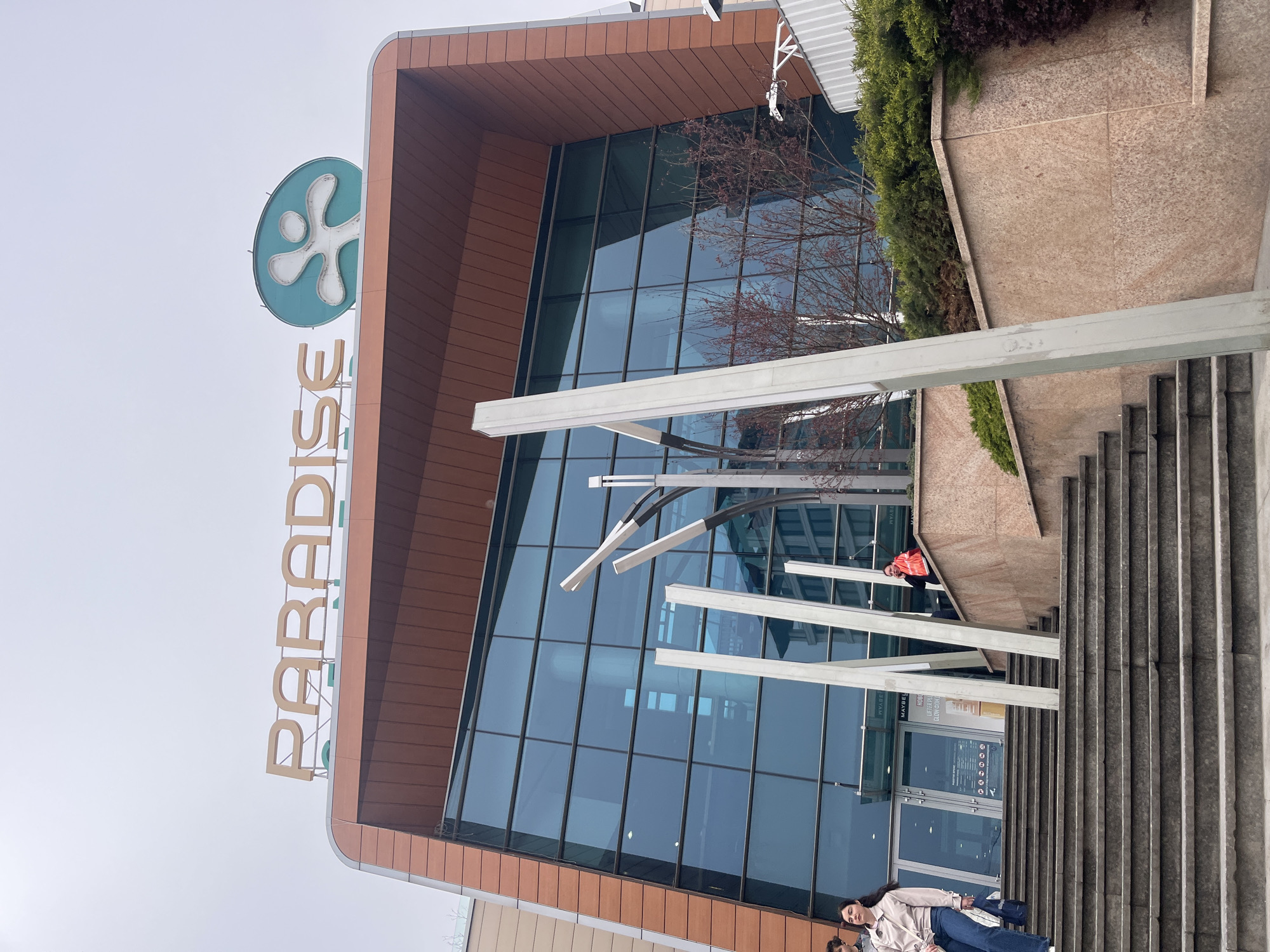 The image depicts the "Paradise" shopping center in Sofia, Bulgaria. The building has a modern design with a large glass facade and a brown exterior. The "PARADISE" sign is vertically oriented on the building's facade. In the foreground, there are concrete steps leading up to the entrance, and modern white architectural elements are visible. Two people are present on the steps: one person is sitting on the stairs, while another is standing. A man is seen partially behind the building, wearing a red vest. The sky is overcast.