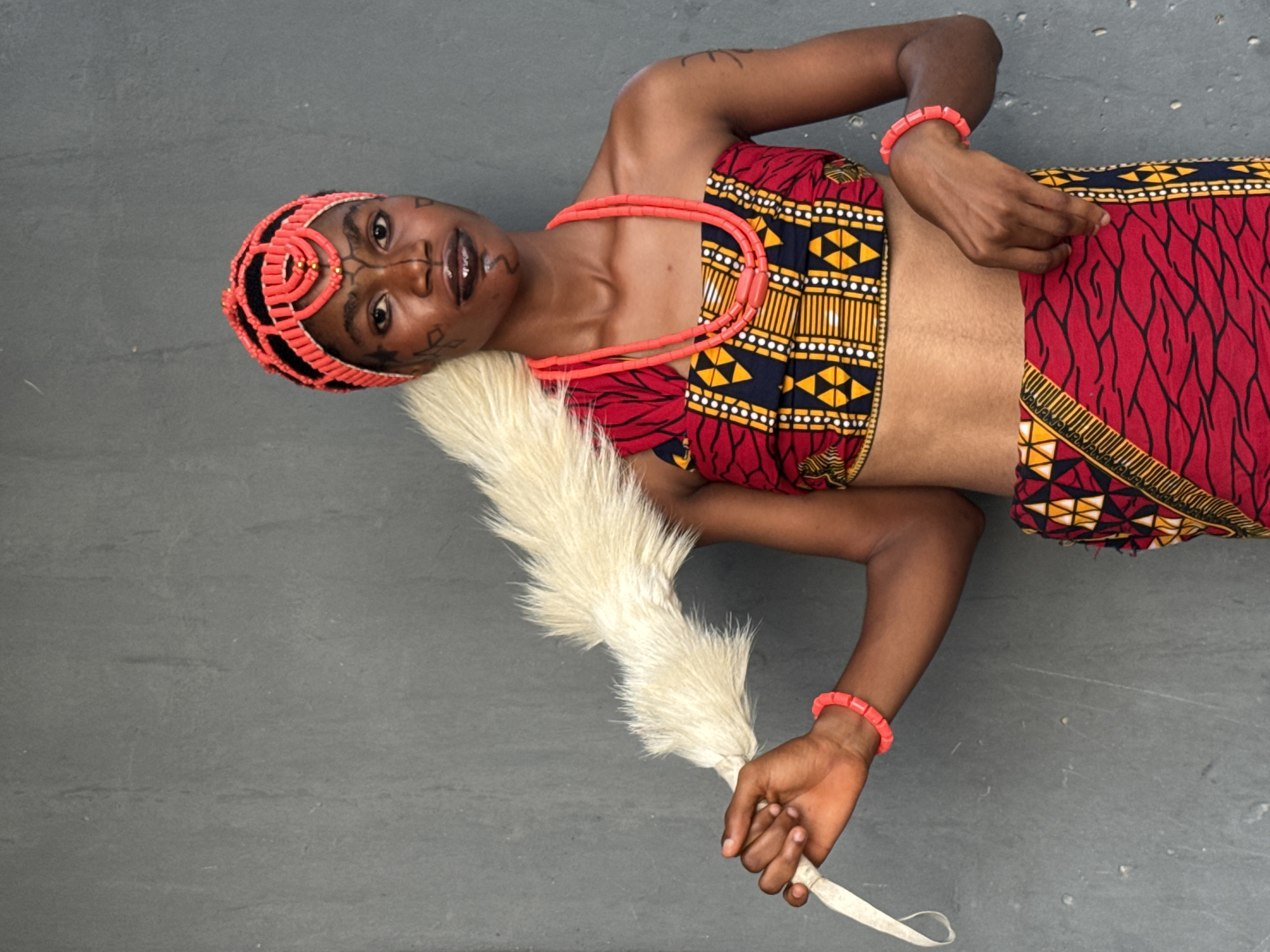 An overhead shot depicts a woman lying on a gray surface. She is dressed in a traditional Igbo attire consisting of a red, yellow, and black patterned top and skirt. She wears a coral bead headdress and necklace, with a coral bead bracelet on each wrist. Her face is adorned with tribal markings. She holds a white feathered fan in her right hand. The location is not evident from the image. No other people or objects are visible.