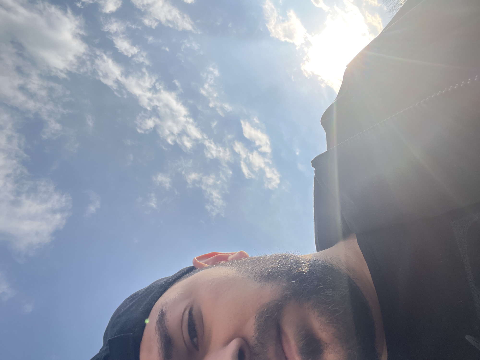A low-angle shot of a man with a beard and black cap, partially obscuring his face. The sky is visible in the background, featuring a mix of blue sky and scattered clouds with the sun shining brightly. No landmarks visible indicate the city of Veliko Tarnovo, Bulgaria.