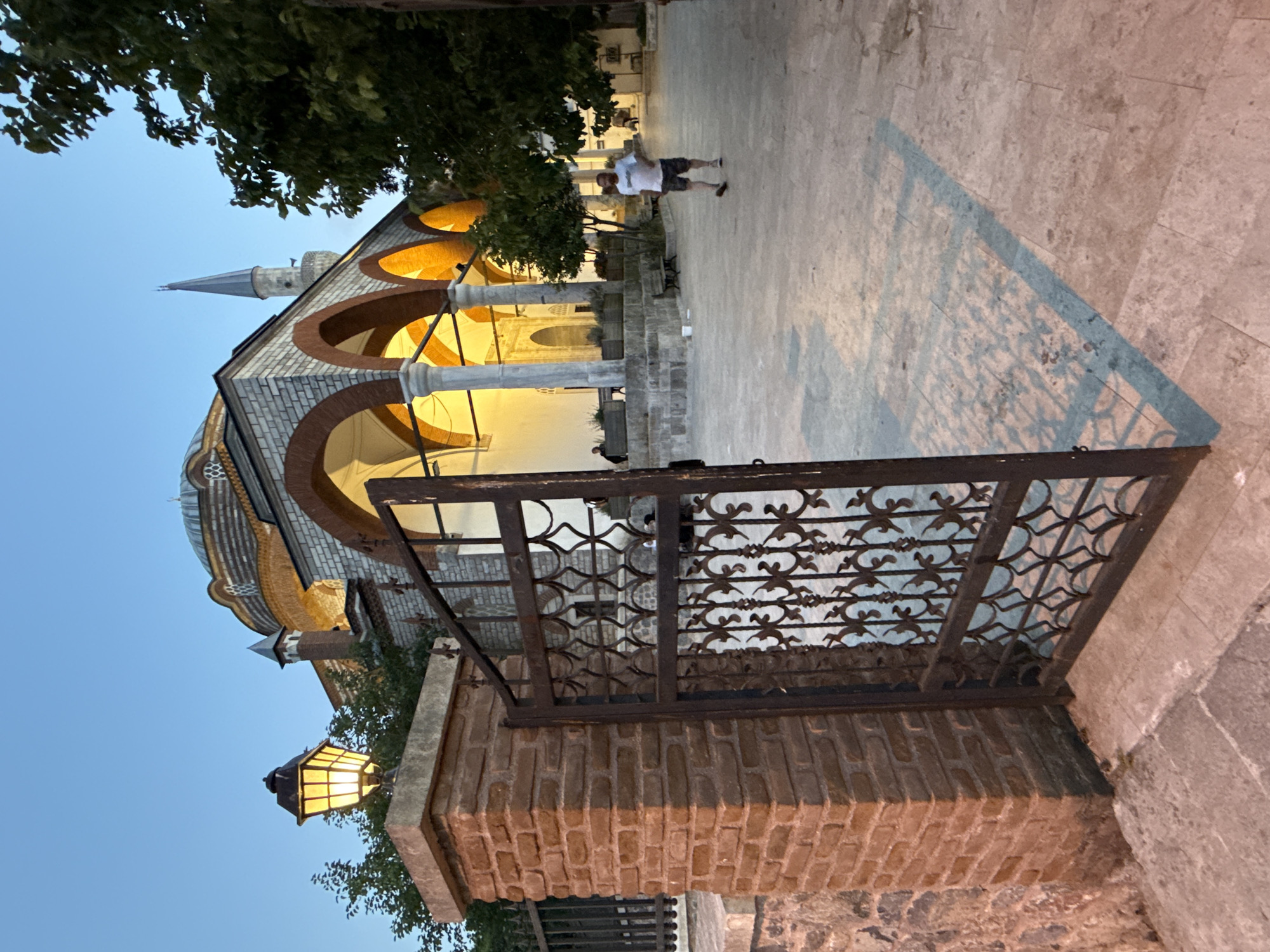 The image shows a view through an open, ornate metal gate towards a mosque complex in Üsküdar, Turkey. The gate is to the left and foreground, made of dark metal with intricate patterns. A brick pillar is to the left of the gate, topped with a lit, antique-style lamp.
Beyond the gate, the mosque features a domed roof and a minaret. The facade has arched walkways supported by columns, illuminated with warm light. The architecture suggests it might be an older, historically significant building. The ground in front of the mosque is paved.
A man in a white t-shirt and dark shorts is walking away from the camera towards the mosque's entrance. Another person is partially visible sitting on a bench in the background. Trees with dark green foliage frame the right side of the scene. The sky is a clear, soft blue, indicating either early morning or late evening. The overall impression is serene and atmospheric.