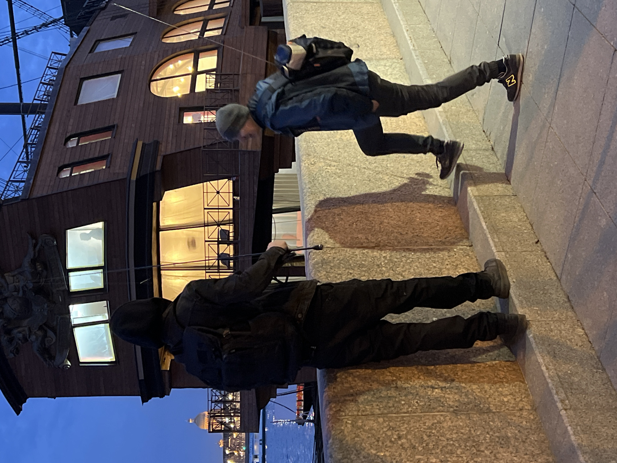 Two people are standing on a granite embankment along the Neva River in Saint Petersburg, Russia. The background features a dark wooden structure with multiple windows, some arched, illuminated from within. In the distance, across the river, is a view of Saint Isaac's Cathedral.
The person closest to the wooden structure is dressed entirely in black, including a hooded jacket and backpack. The other individual wears a blue jacket, black pants, a gray beanie, and New Balance sneakers. They are reaching out with a long pole in their hand, seemingly interacting with the person in black.