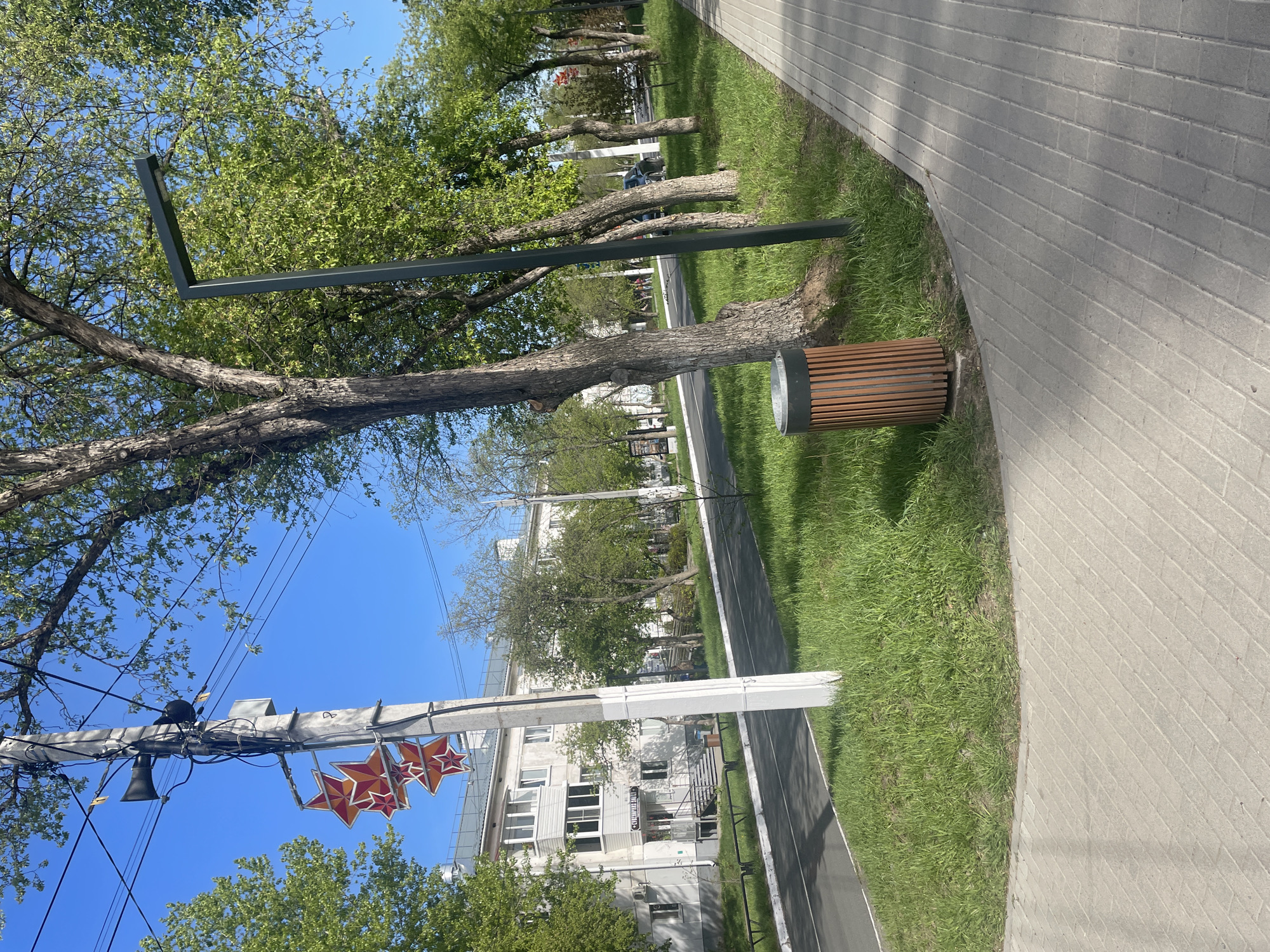 The image shows a street scene in Chaikovsky, Russia. The view is oriented parallel to a paved walkway with a grassy area between it and a roadway. Along the walkway, there are trees, a modern streetlight, and a wood-slatted waste bin. A white utility pole is visible with a speaker and three orange and red five-pointed stars attached. In the background, there is a multi-story building. The sky is clear and blue.