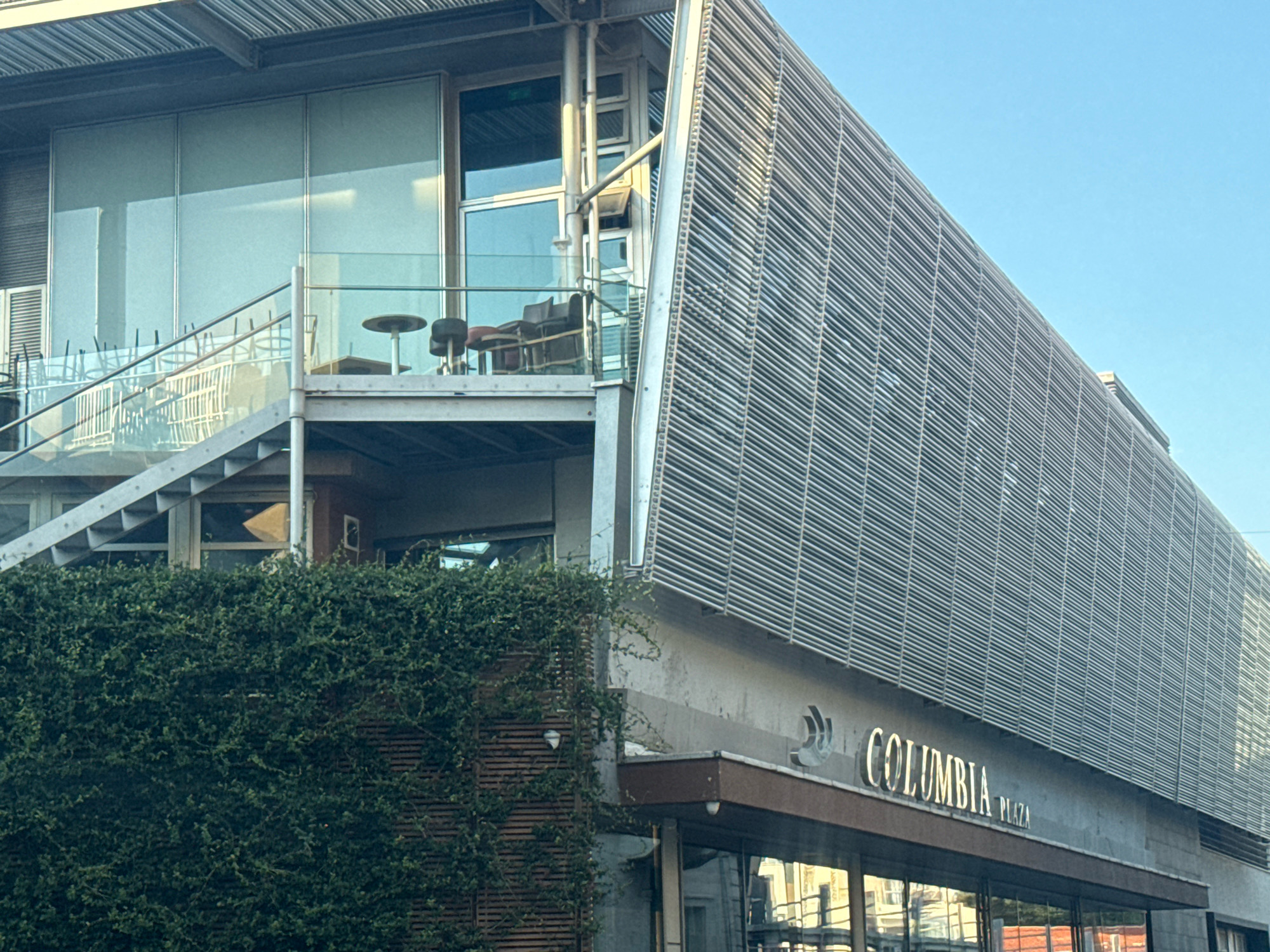 The image shows the facade of a modern building in Limassol, Cyprus, with a prominent "COLUMBIA PLAZA" sign. The building features a distinctive slatted exterior made of metal, creating a textured surface. A green ivy-covered wall extends up the side of the building on the left. Above the ivy, there is an outdoor seating area with tables and chairs visible through a glass facade. A metal staircase with glass railings leads up to this seating area. The sky above is clear and blue. The overall impression is one of a contemporary urban structure, possibly a commercial or residential complex.
