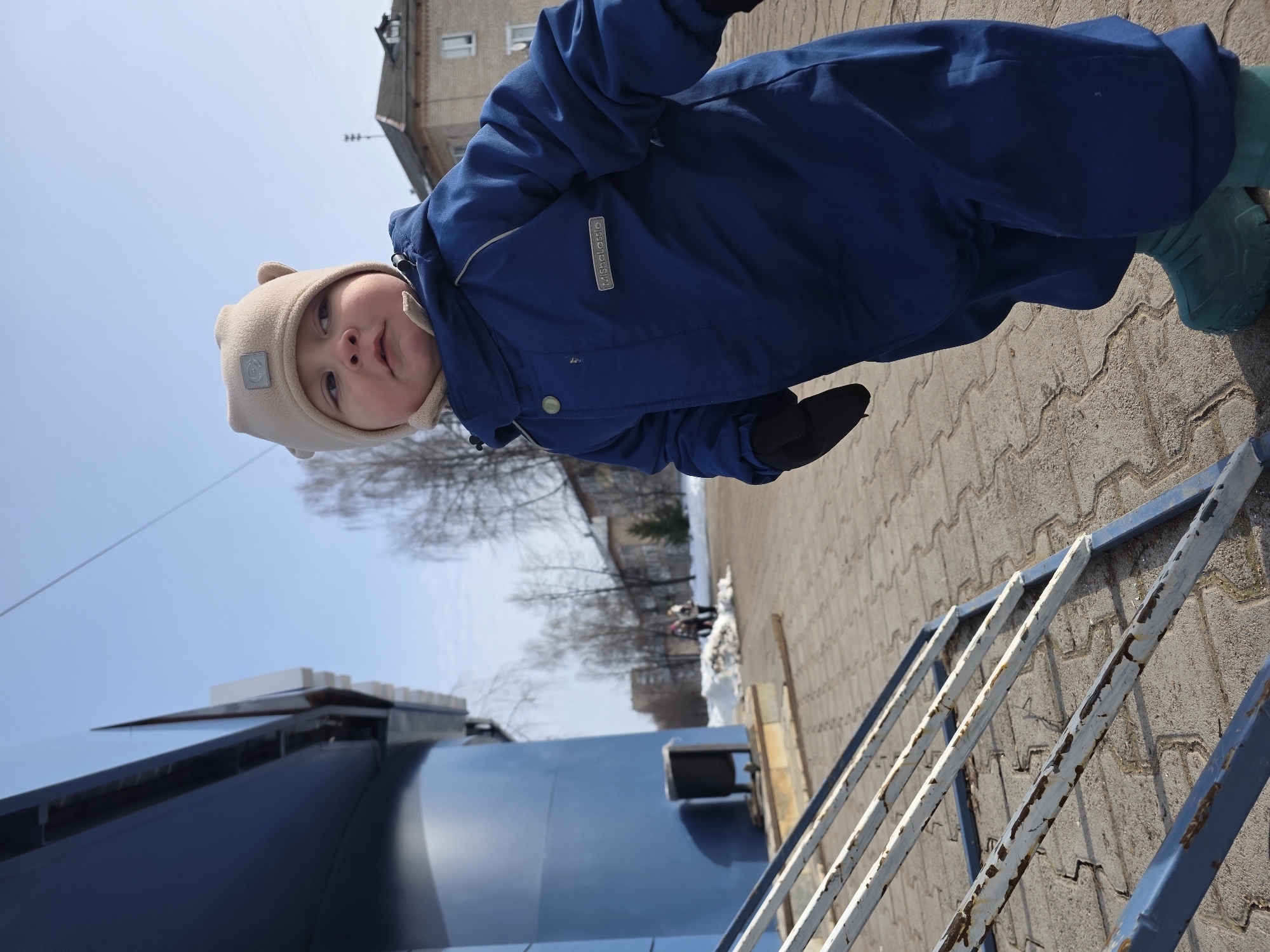The scene depicts a young child standing on a paved walkway in the city of Chaikovsky, Russia. The child, a toddler, is dressed in a dark blue insulated jumpsuit with a "MishaLassie" brand tag on the left chest, a light beige beanie with ear flaps, black gloves, and teal boots. The child's face is visible, looking to the upper right, with light blue eyes.
To the immediate lower left, a weathered metal structure with peeling white paint over blue, revealing rust, lies on the ground. The walkway itself is paved with interlocking light-colored concrete blocks. A large, modern-style building with dark blue panels and curved sections is prominent on the left side of the frame. A black cylindrical trash receptacle stands near the base of this building.
In the background, under a clear blue sky, bare deciduous trees and patches of dirty snow are visible on the ground. Further back, several traditional multi-story brick buildings with dark sloped roofs can be seen, typical of urban architecture. A small antenna is visible on the roof of one distant building. Several out-of-focus individuals are discernible walking in the mid-distance near the snow patches. The lighting indicates a bright, sunny day.